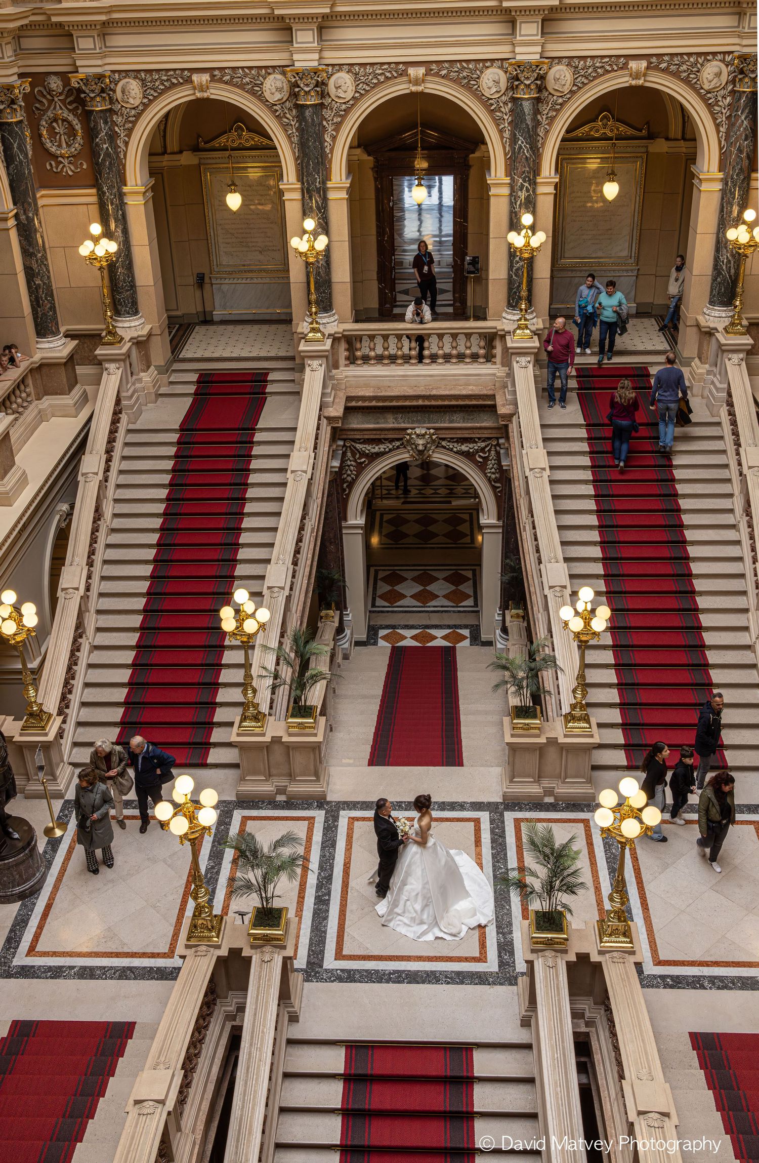 Aerial view of elegant wedding couple on ornate marble staircase with red carpet in grand historic building.