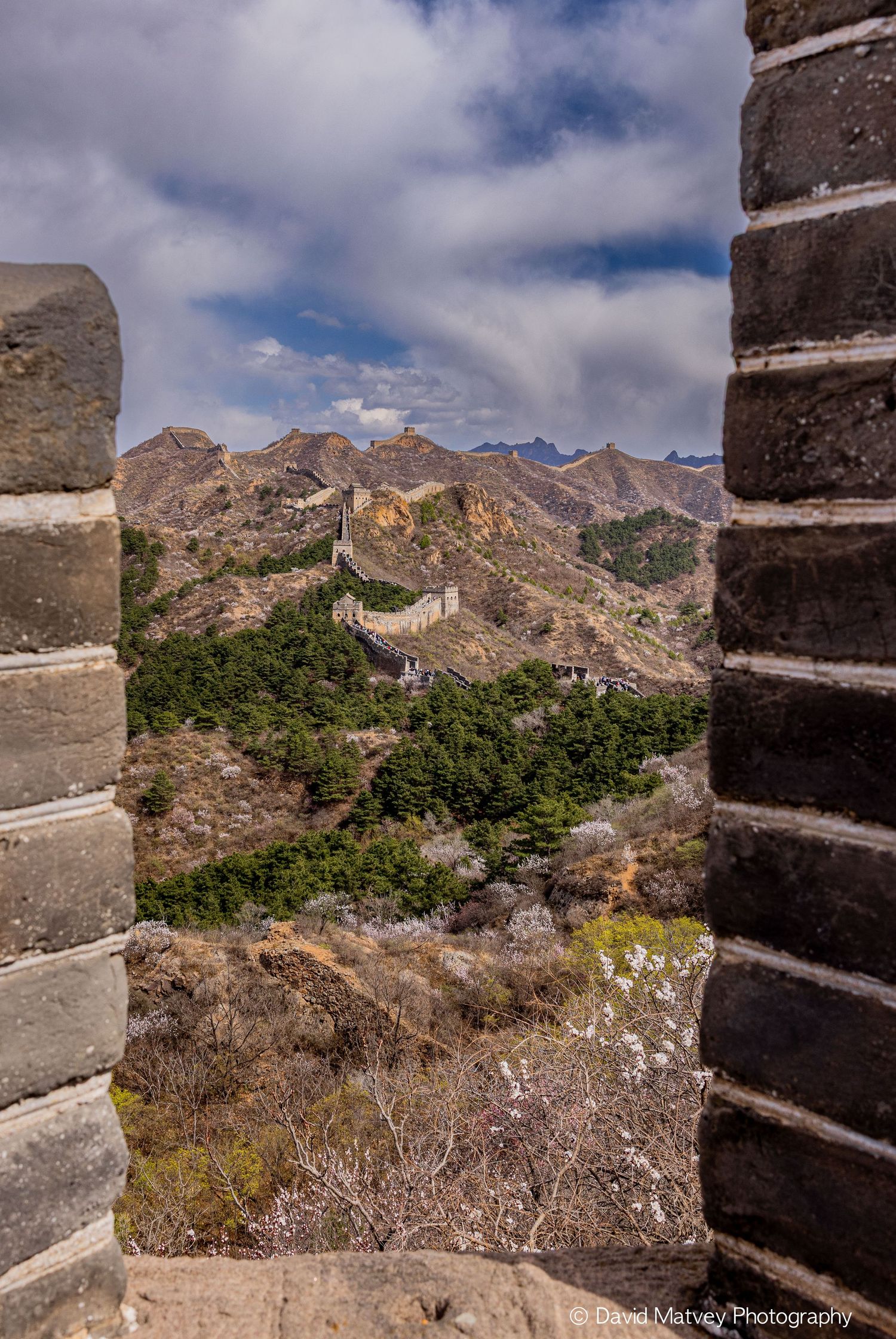 Great Wall of China stretching across rugged mountain ridges viewed through stone tower opening under cloudy sky.