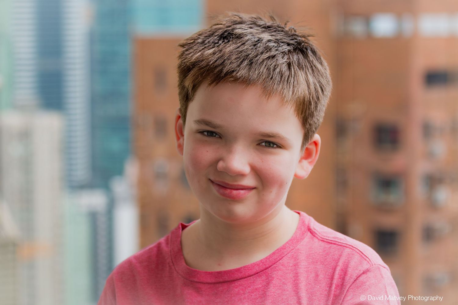 Young boy in pink shirt smiling with city buildings in the background.