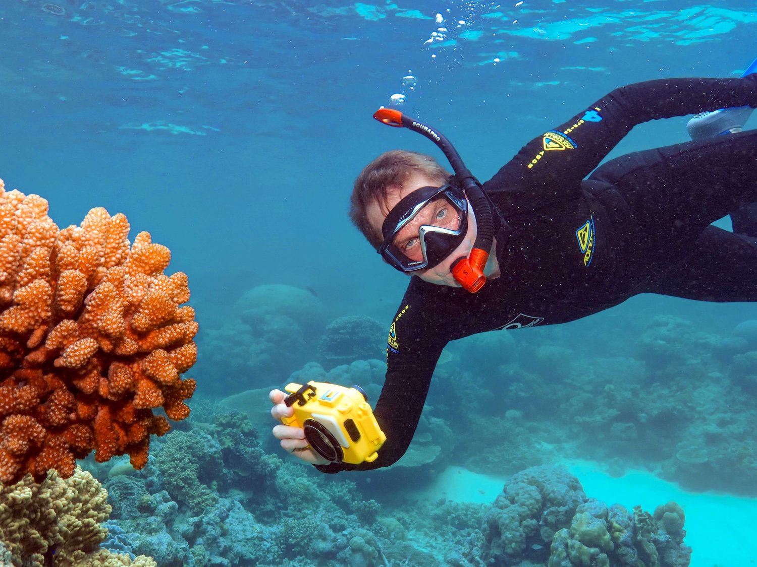 Smiling snorkeler in black wetsuit holds yellow camera near colorful coral reef in clear blue tropical water.
