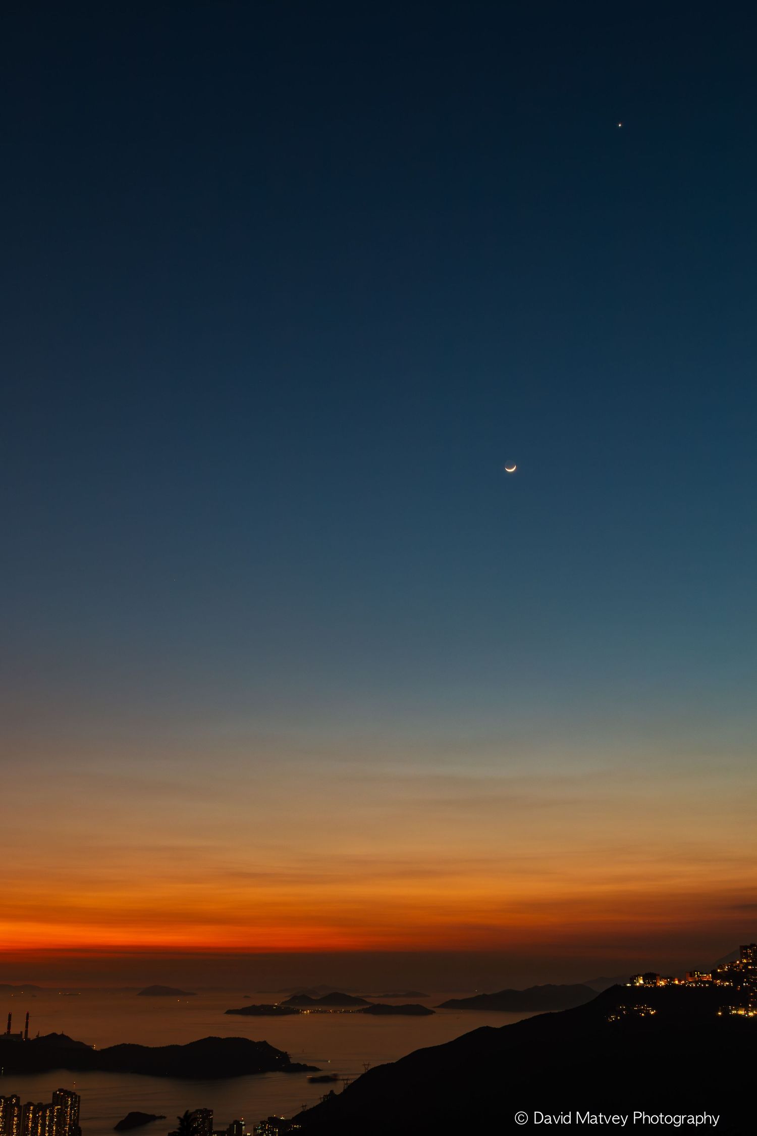 Crescent moon and bright star above vibrant orange and blue sunset over coastal city skyline.