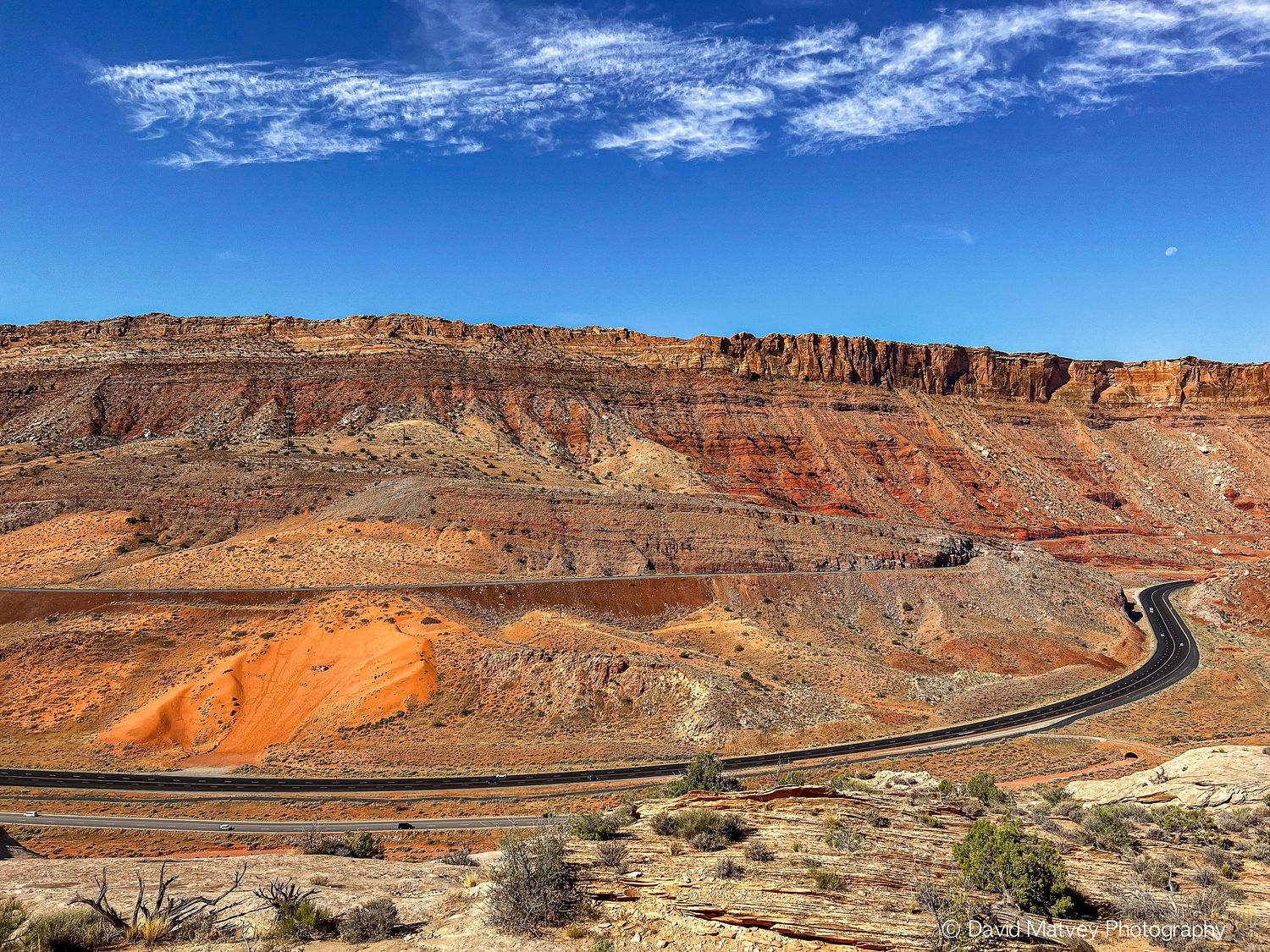Scenic winding highway through layered red sandstone cliffs and desert landscape under clear blue sky.