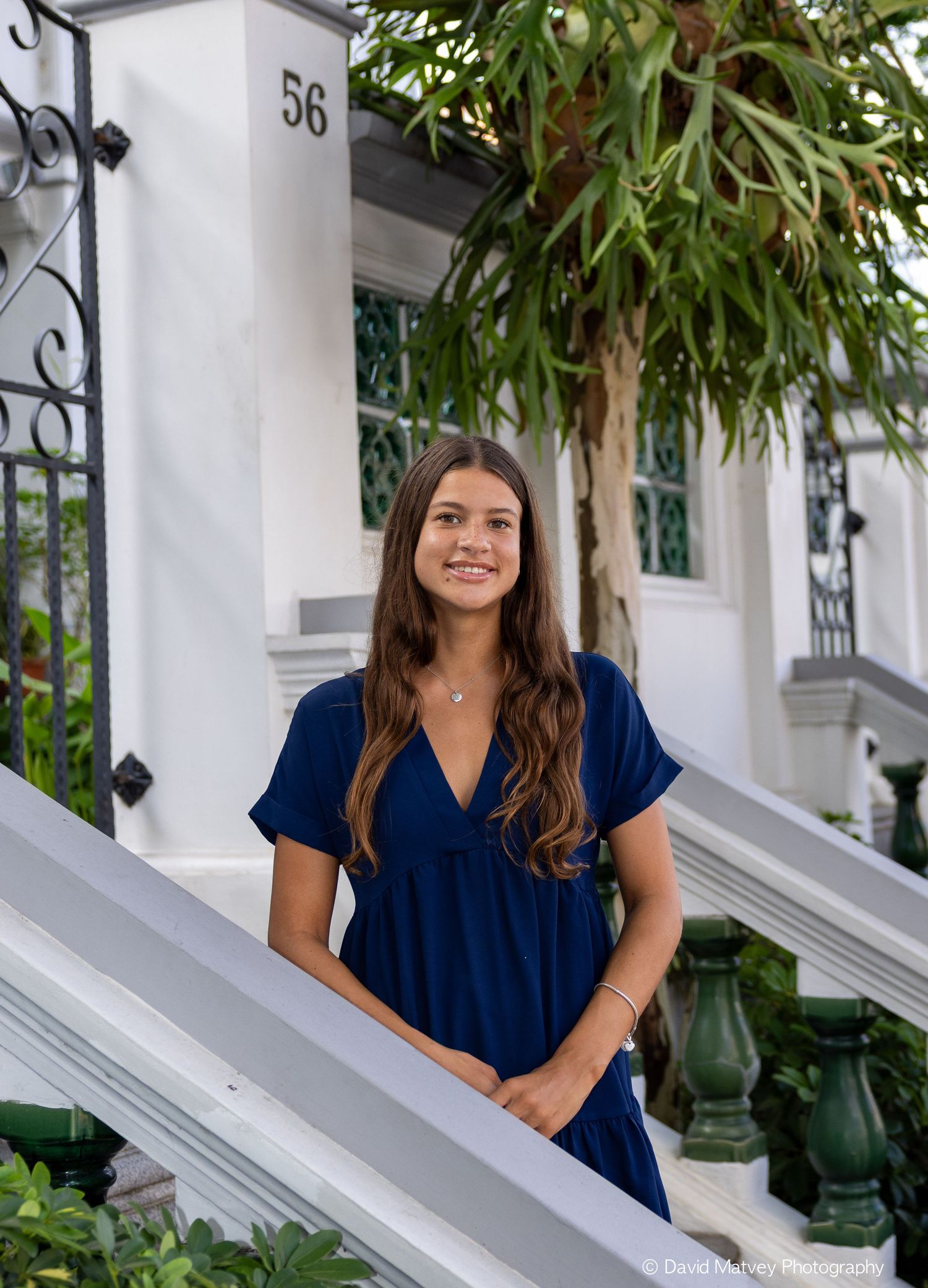 Teenage girl in navy blue dress standing on white steps near lush greenery.