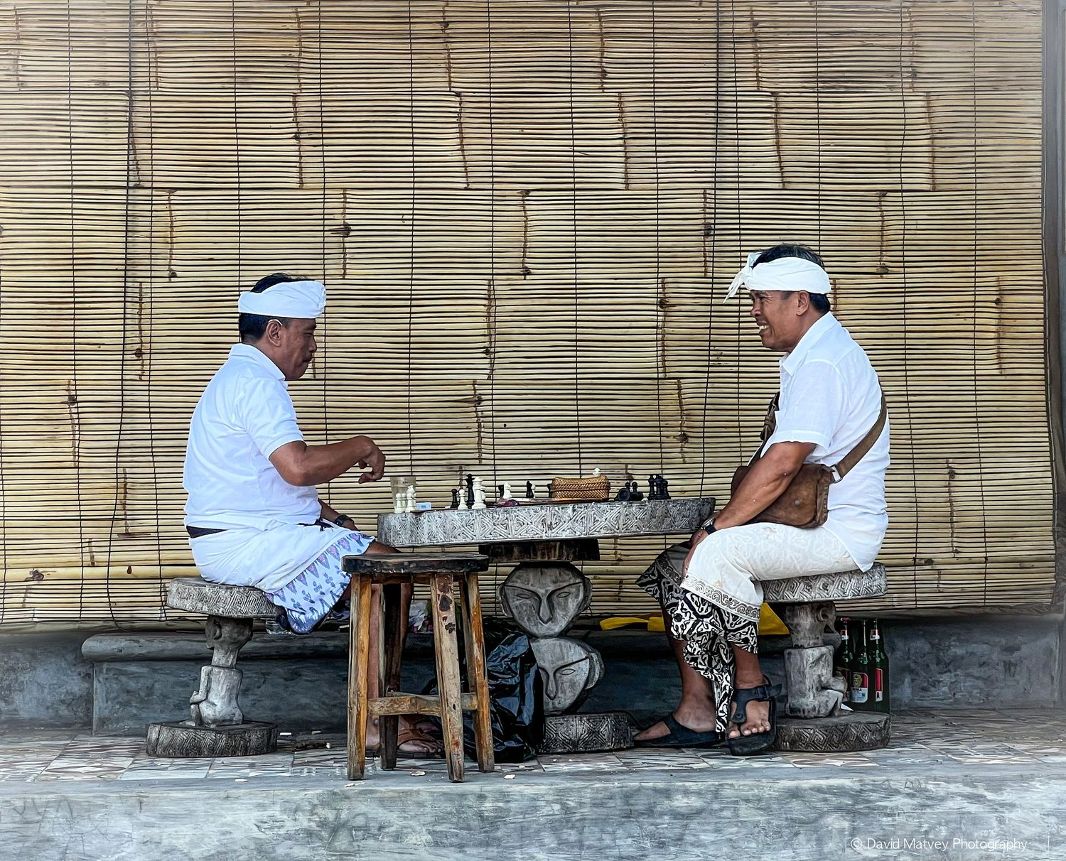 Two Balinese men in white clothing playing chess outdoors.