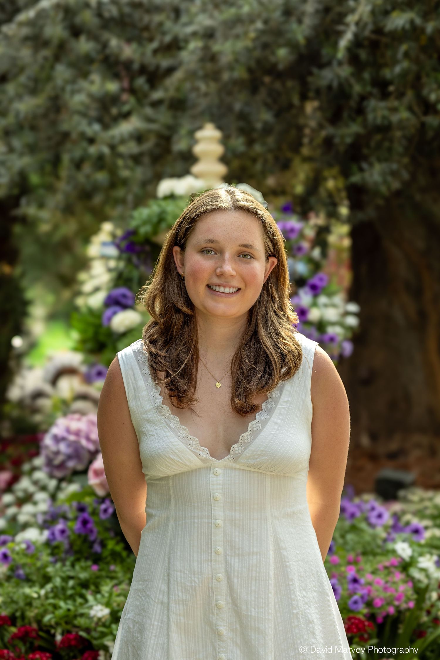 Young woman in white sundress smiling in a colorful garden with purple and white flowers.