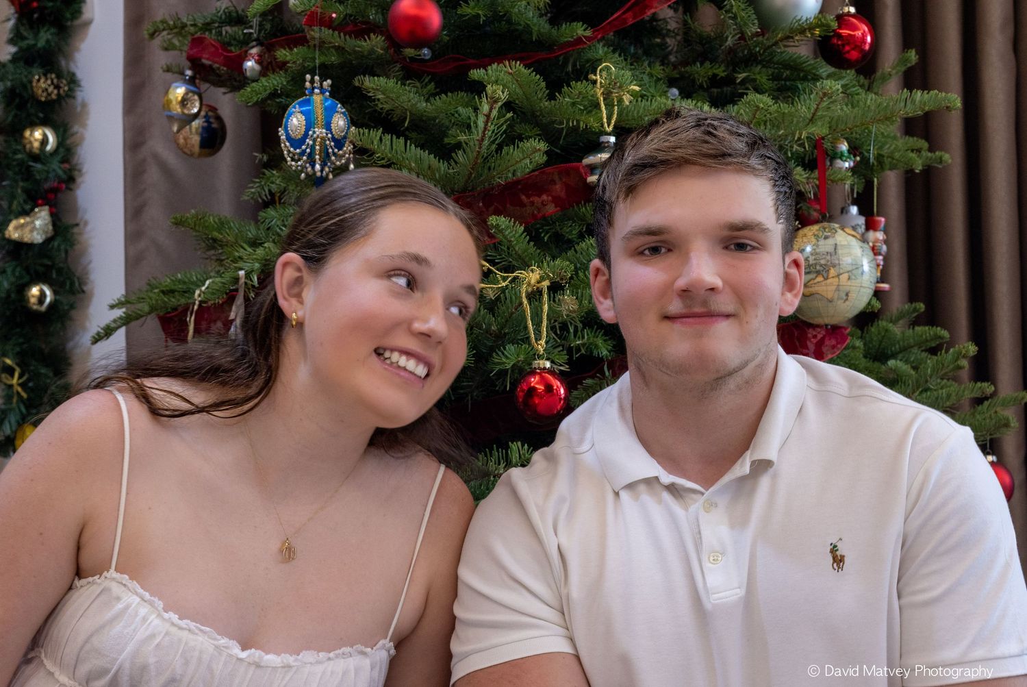 Young smiling couple posing together in front of decorated Christmas tree with red ornaments.