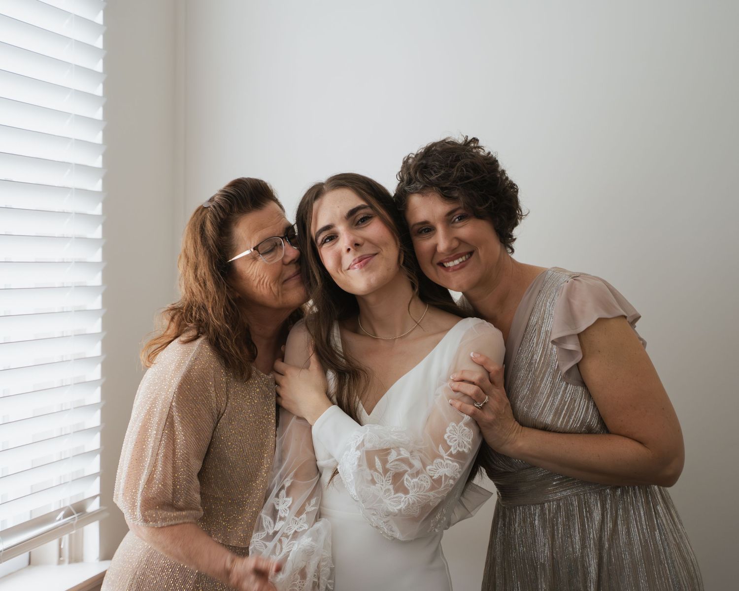 Bride in lace wedding gown posing warmly with two women in a bright, window-lit indoor setting.