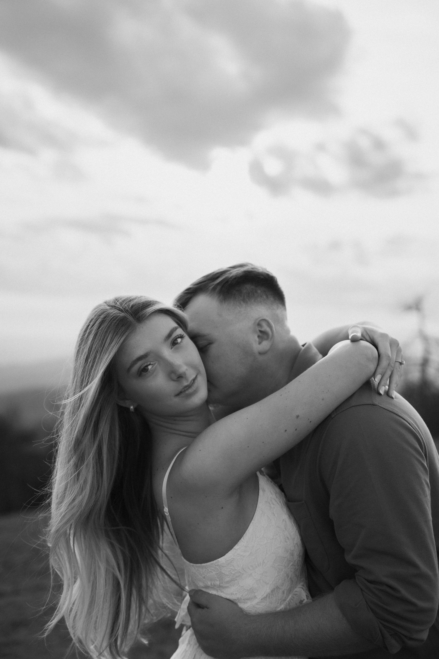 Black and white photo of couple embracing, man kissing woman's cheek against cloudy sky.