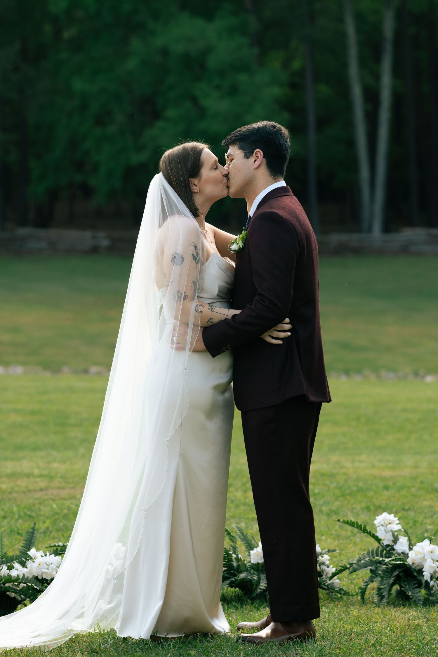 Bride in white gown and groom in burgundy suit share first kiss at outdoor garden wedding ceremony.