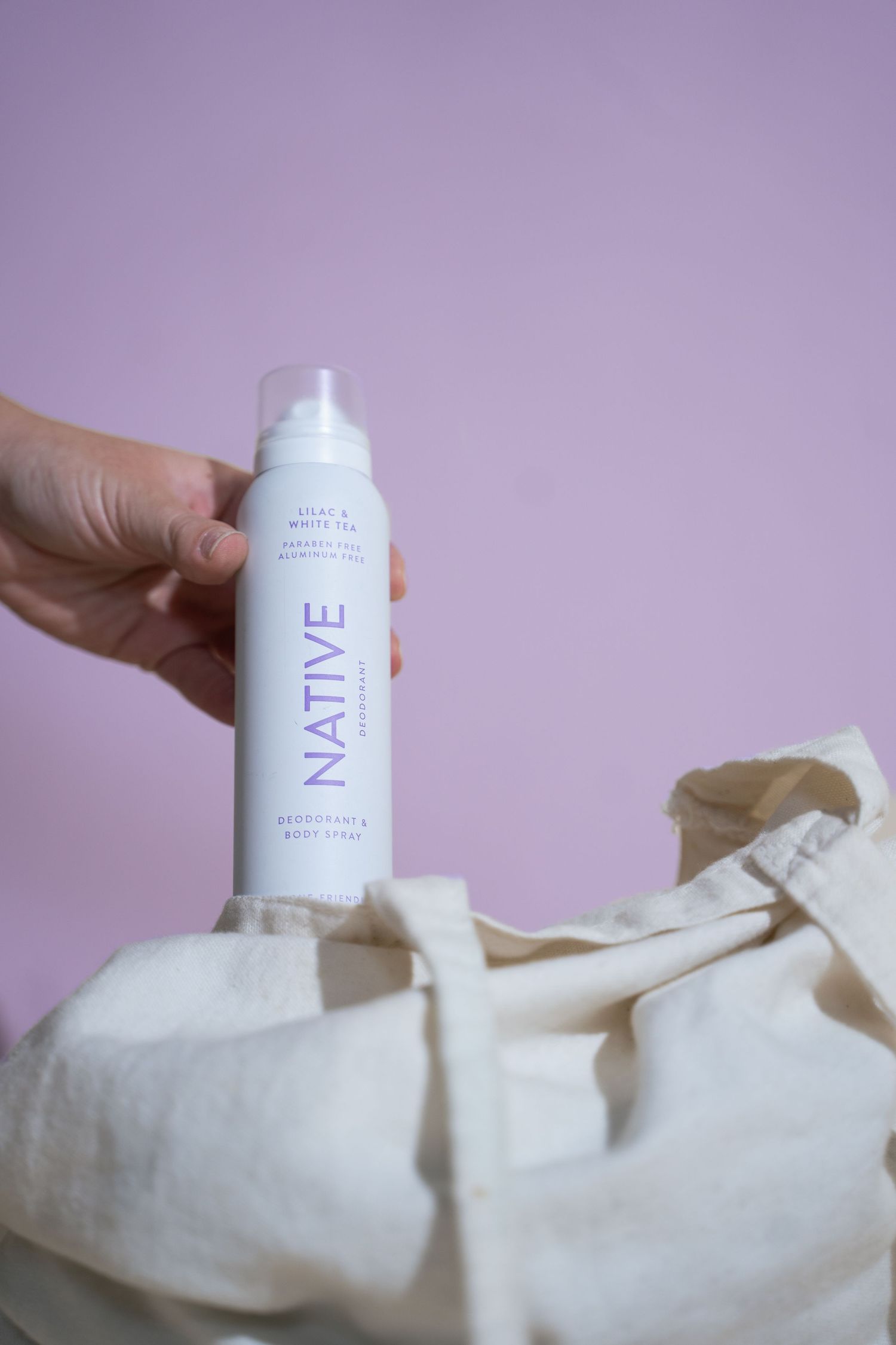 Hand placing Native lilac white tea deodorant body spray into a canvas tote bag on pink background.