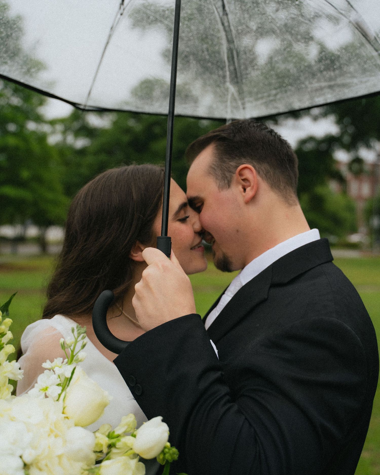 Bride and groom sharing a romantic moment under a clear umbrella in the rain, bride holding white bouquet.
