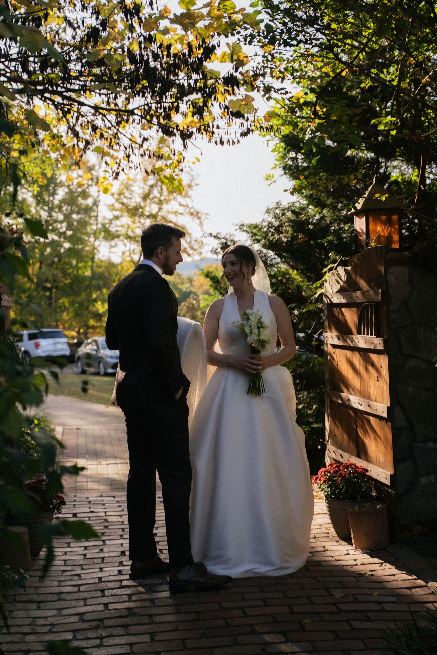 Bride and groom standing on brick pathway surrounded by lush greenery and autumn foliage at golden hour.