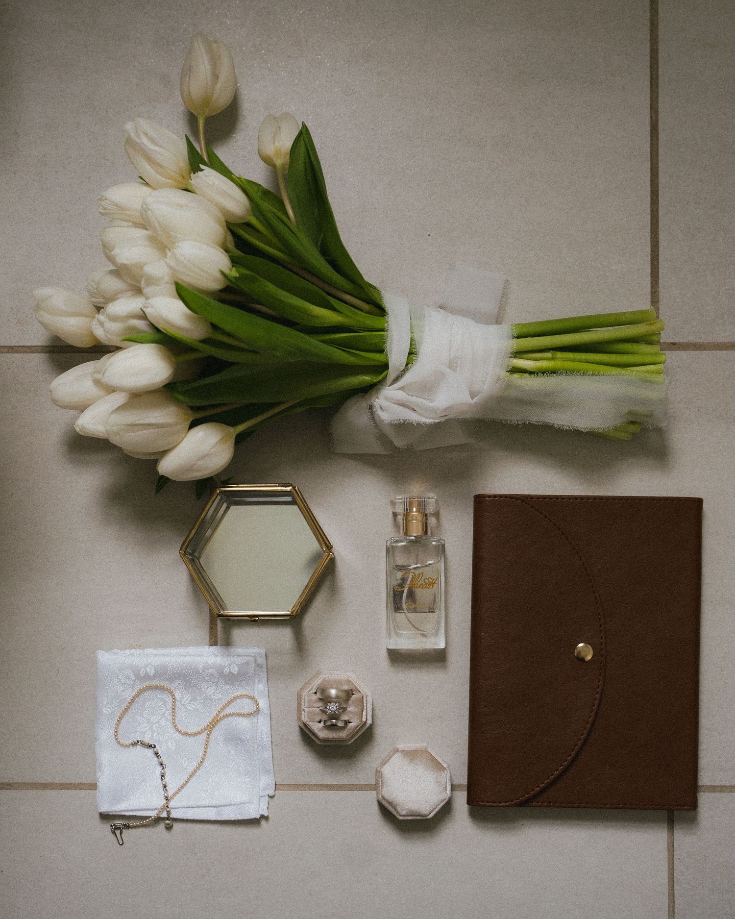 Flat lay of white tulip bouquet, hexagonal mirror, perfume bottle, ring box, necklace, and brown leather journal on beige tile.