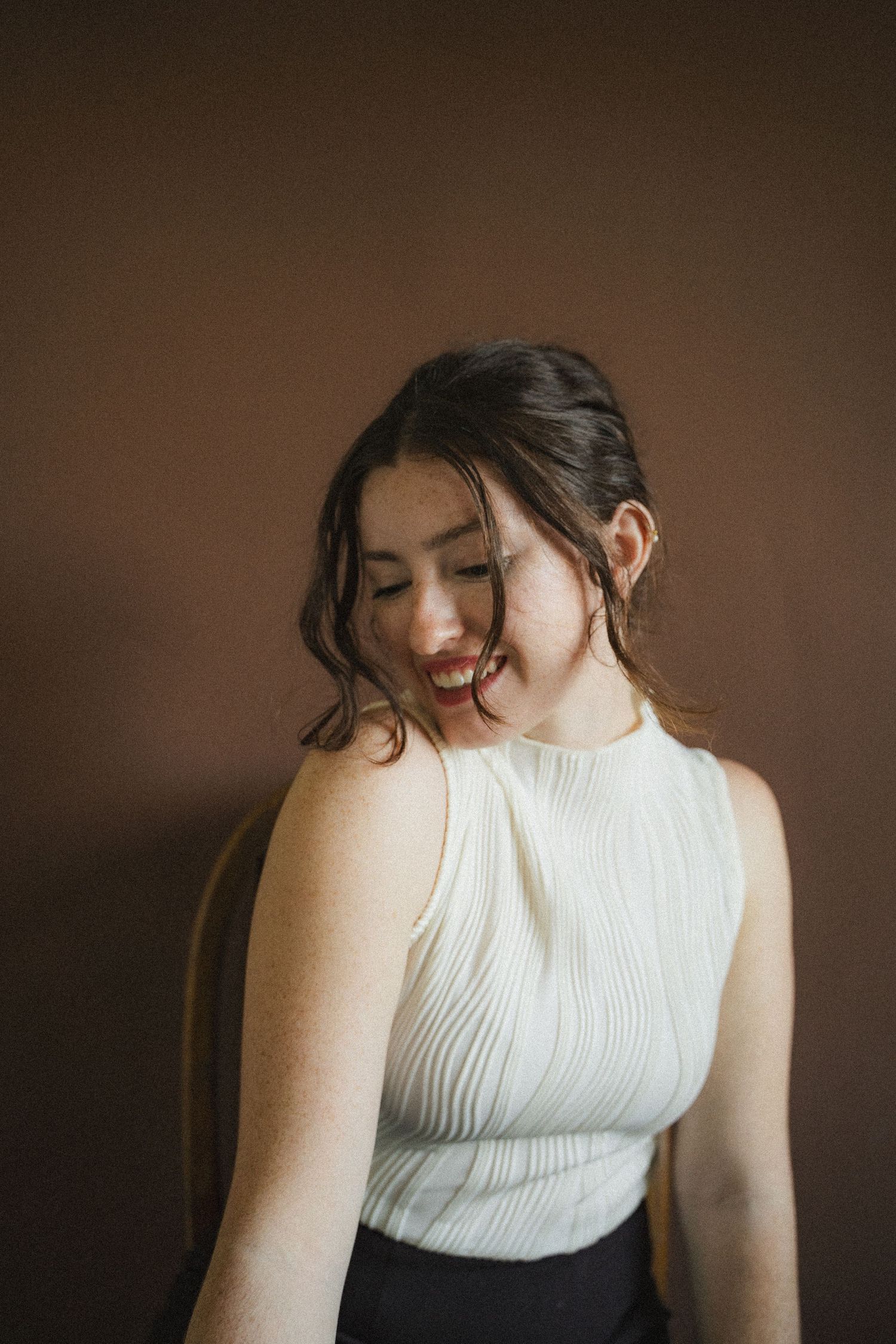 Young woman in a white pleated sleeveless top smiling downward against a warm brown background.