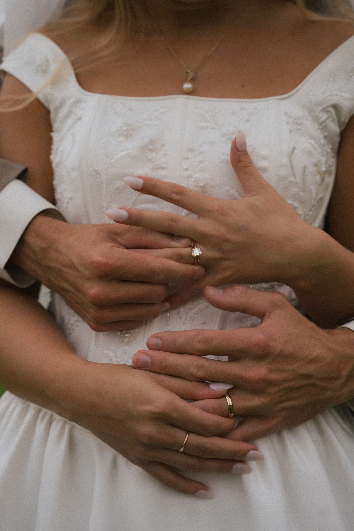 Close-up of newlyweds hands showing diamond engagement ring and gold wedding bands against white embroidered bridal gown.