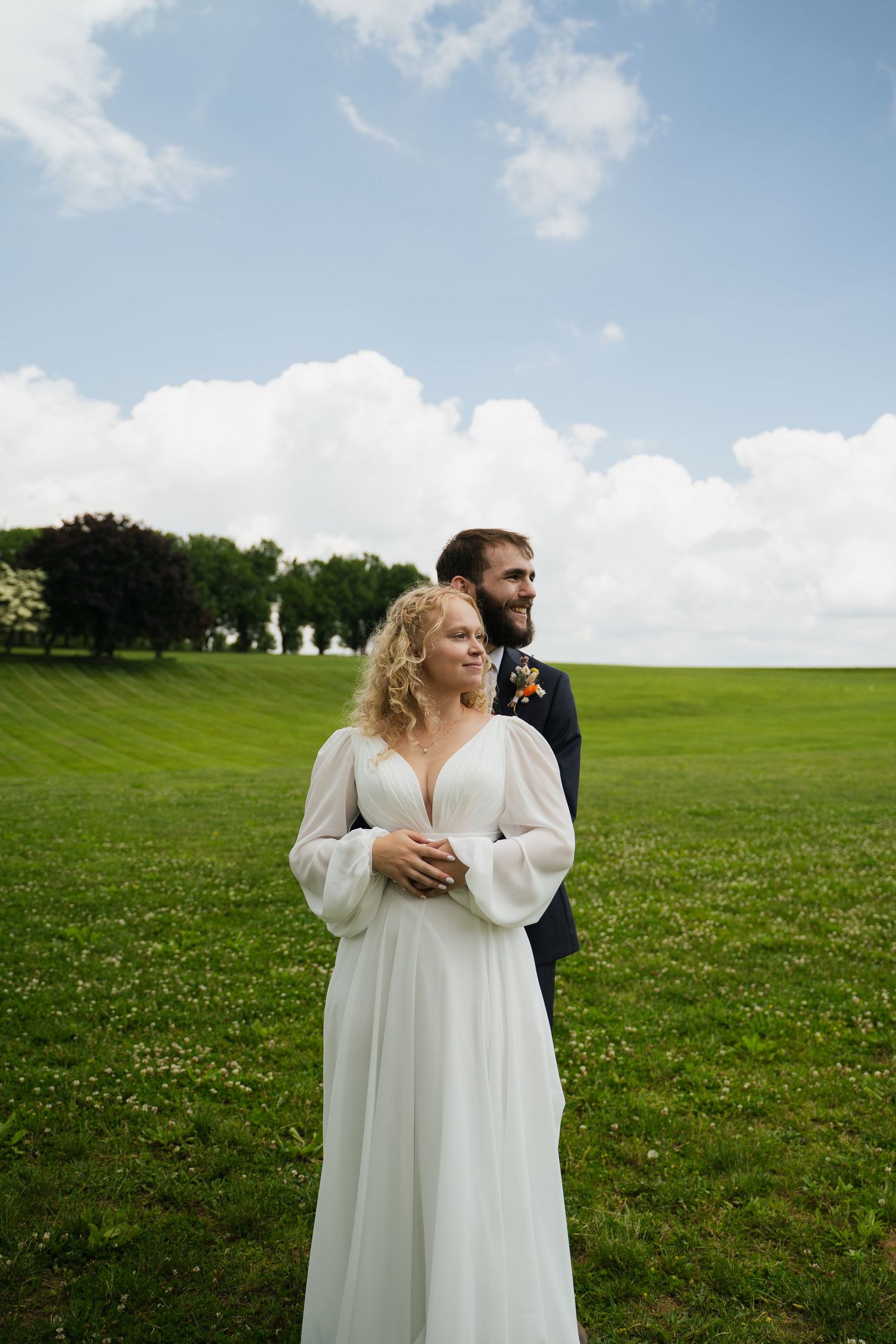 Bride in flowing white long-sleeve gown stands with groom in navy suit on lush green field under cloudy sky.