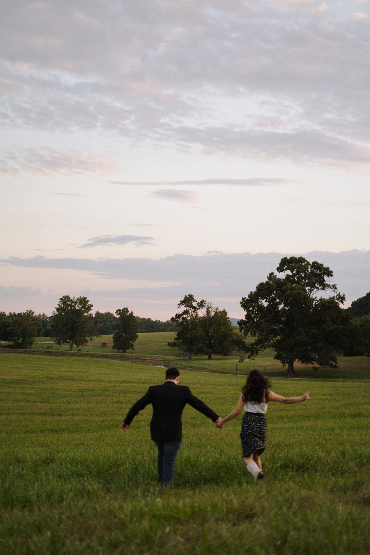 Couple holding hands walking through a vast green field at dusk under a pastel cloudy sky.
