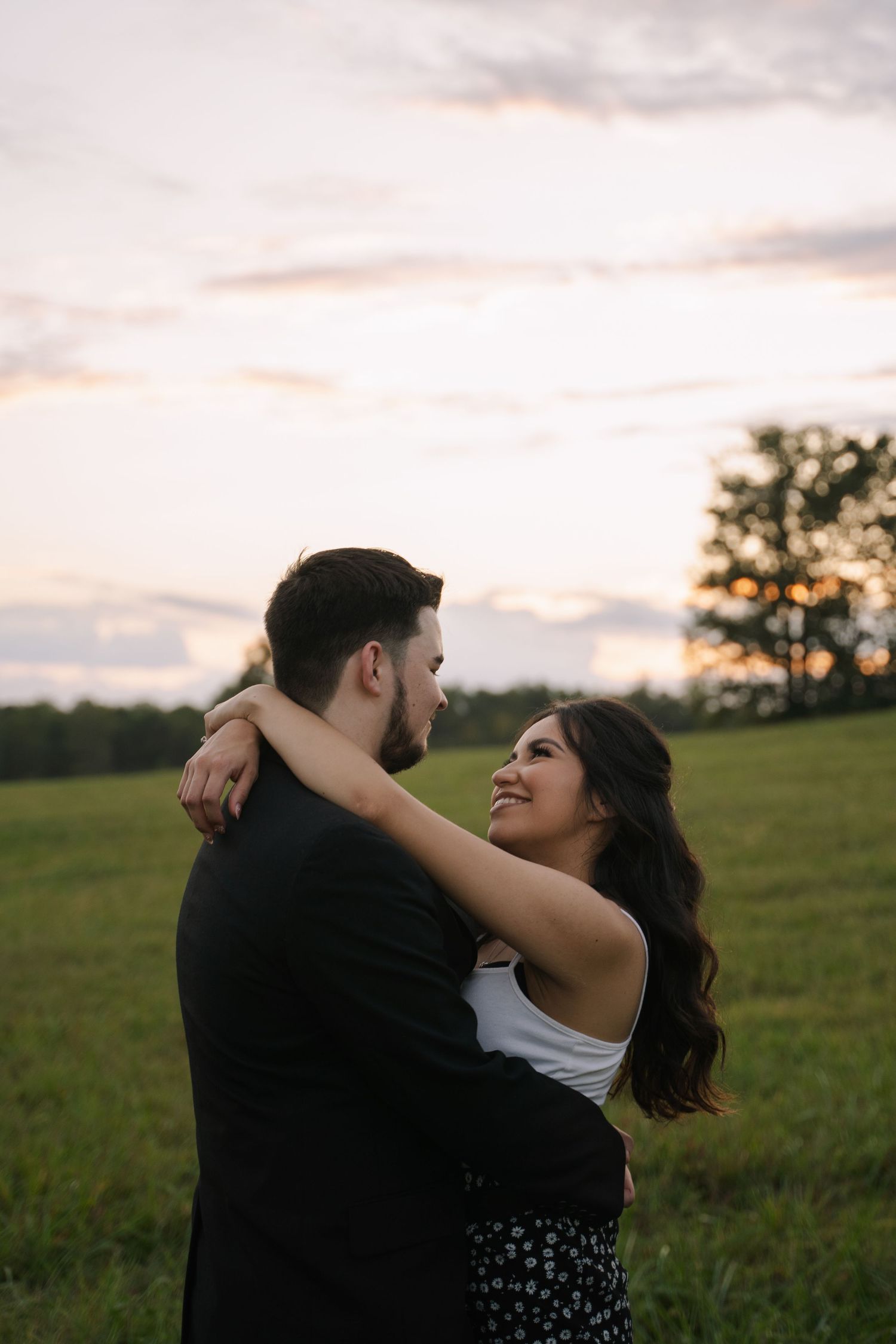 Couple embracing in a green field at sunset, woman smiling up at man in black jacket.