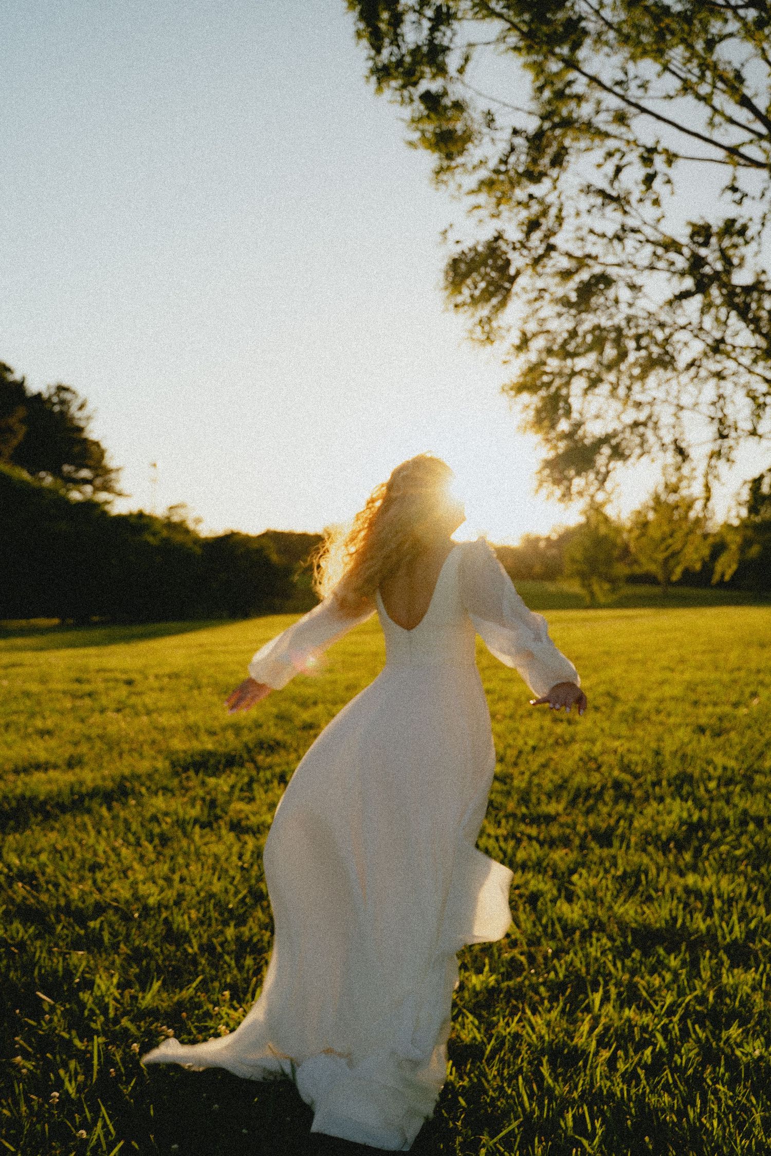 Bride in flowing white long-sleeve gown twirling in sunlit green meadow during golden hour sunset.