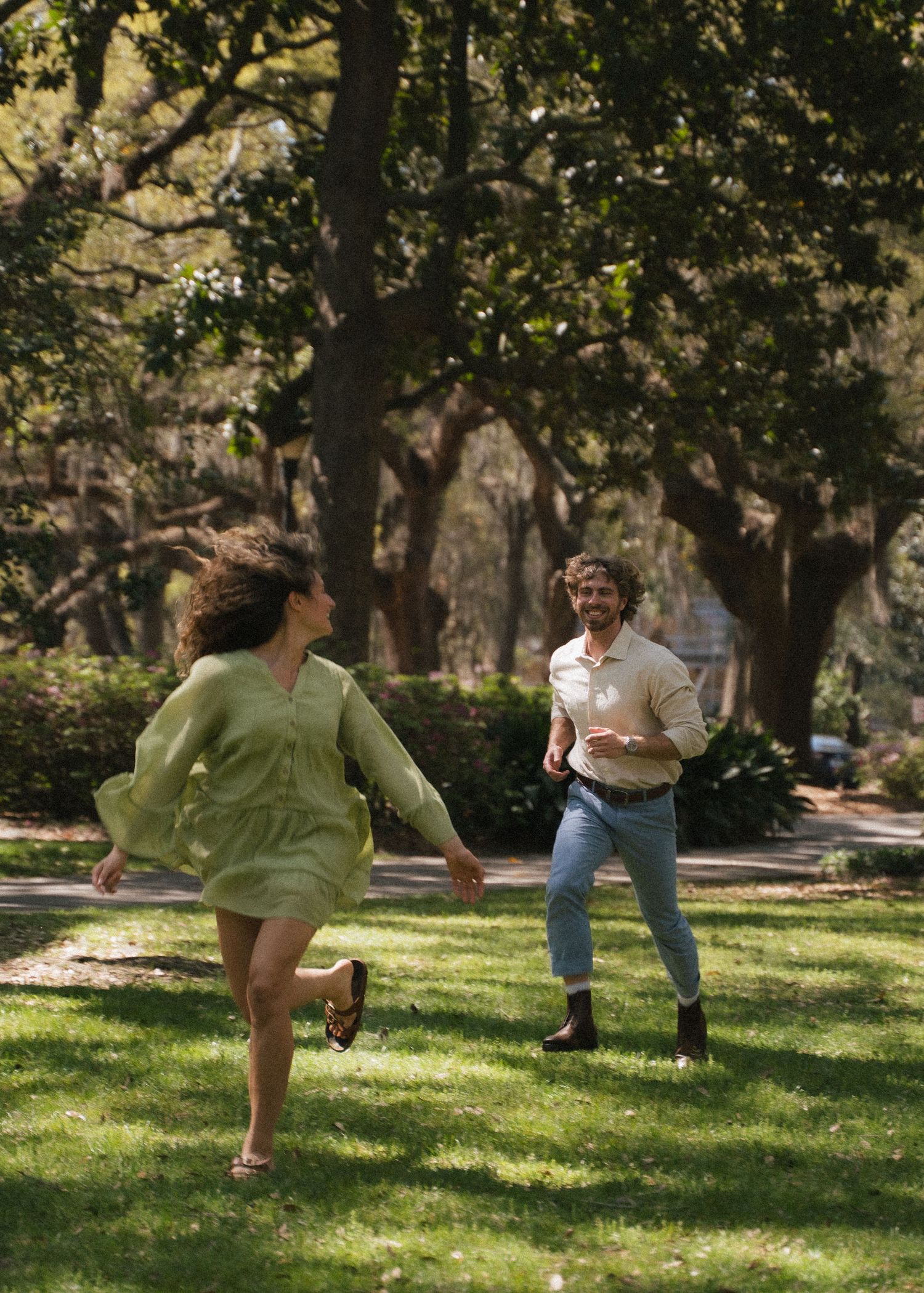 Joyful couple running and holding hands through sunlit park during engagement shoot.