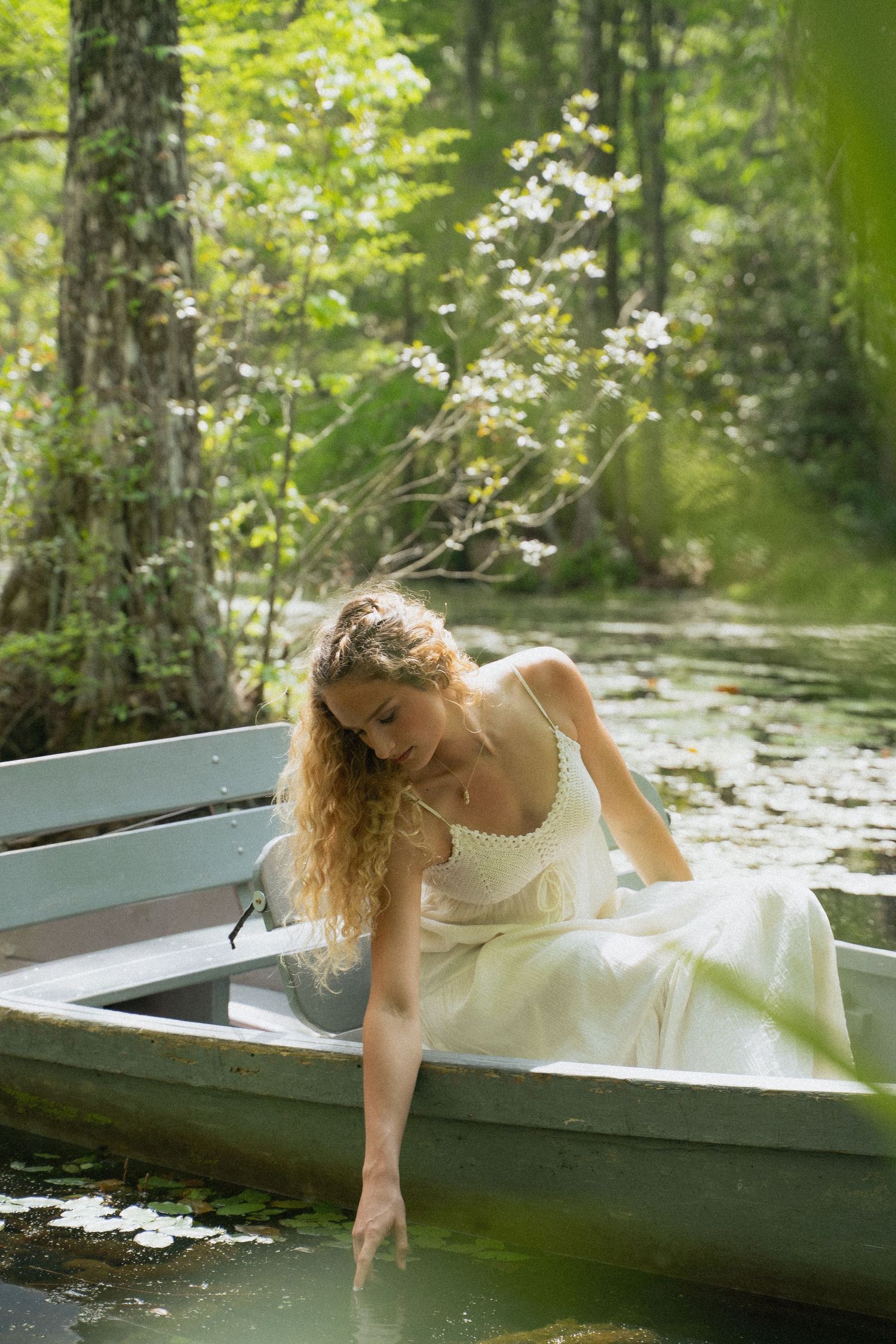 Bride in white gown sits in rowboat trailing fingers in water surrounded by lush green forest.