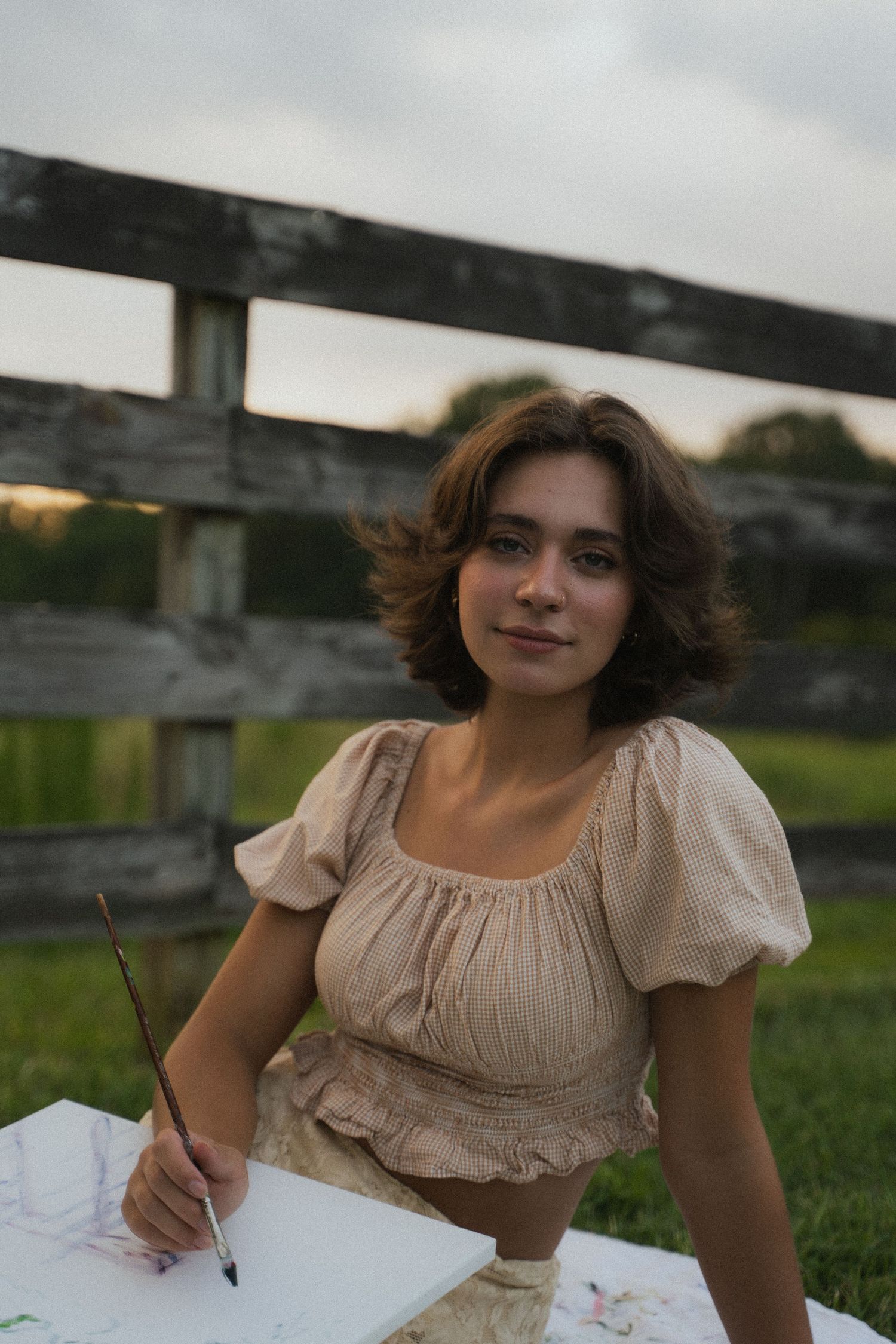 Young woman in beige blouse painting outdoors near a wooden fence at dusk.