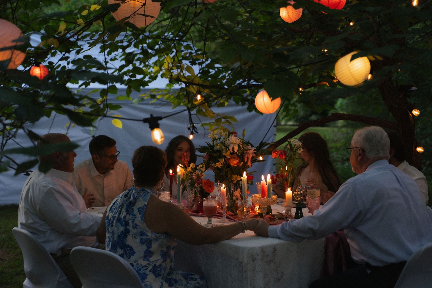 Wedding guests gathered around candlelit outdoor dinner table under colorful hanging lanterns.