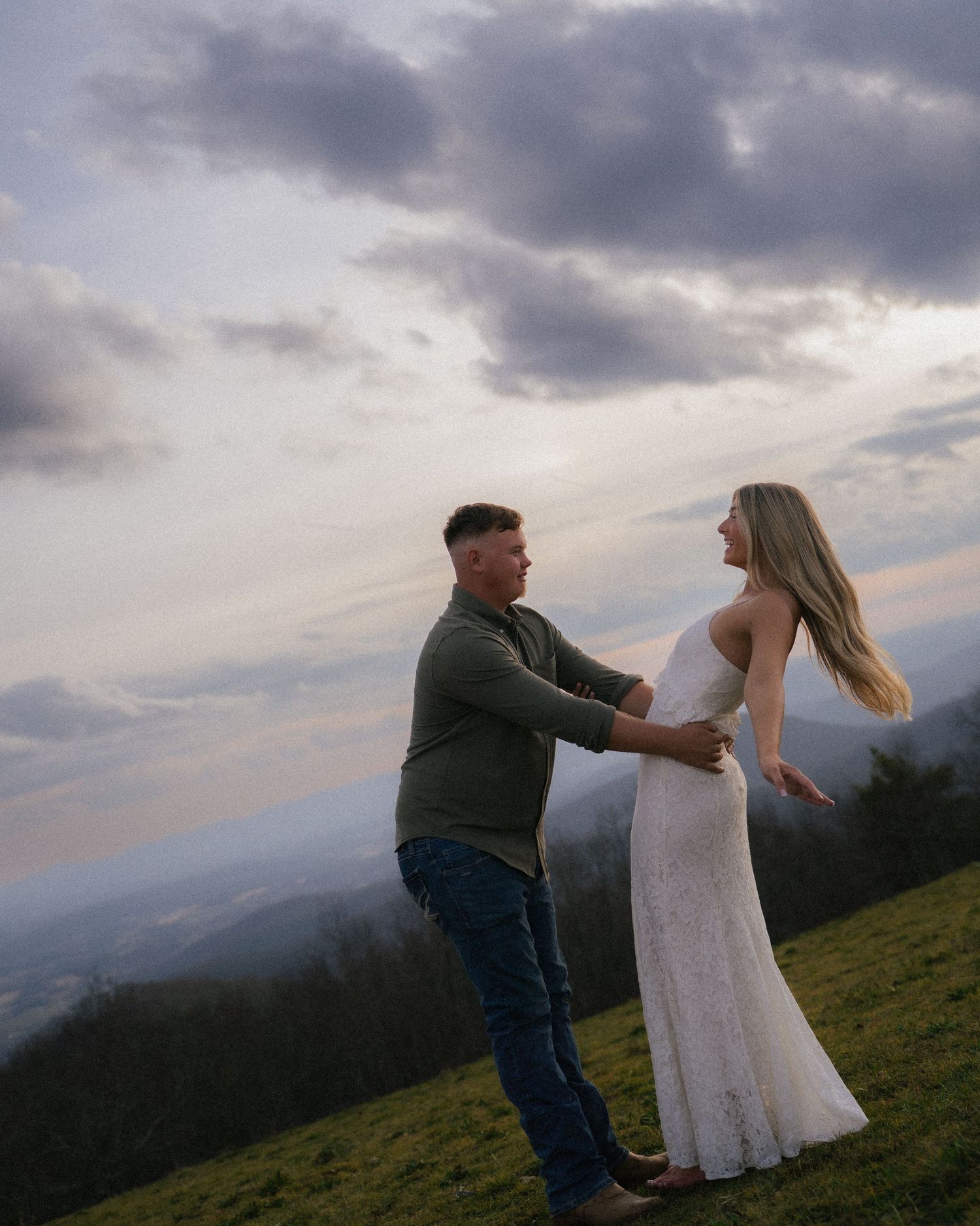 Couple dancing together on a hilltop at sunset with dramatic cloudy sky and mountain valley backdrop.