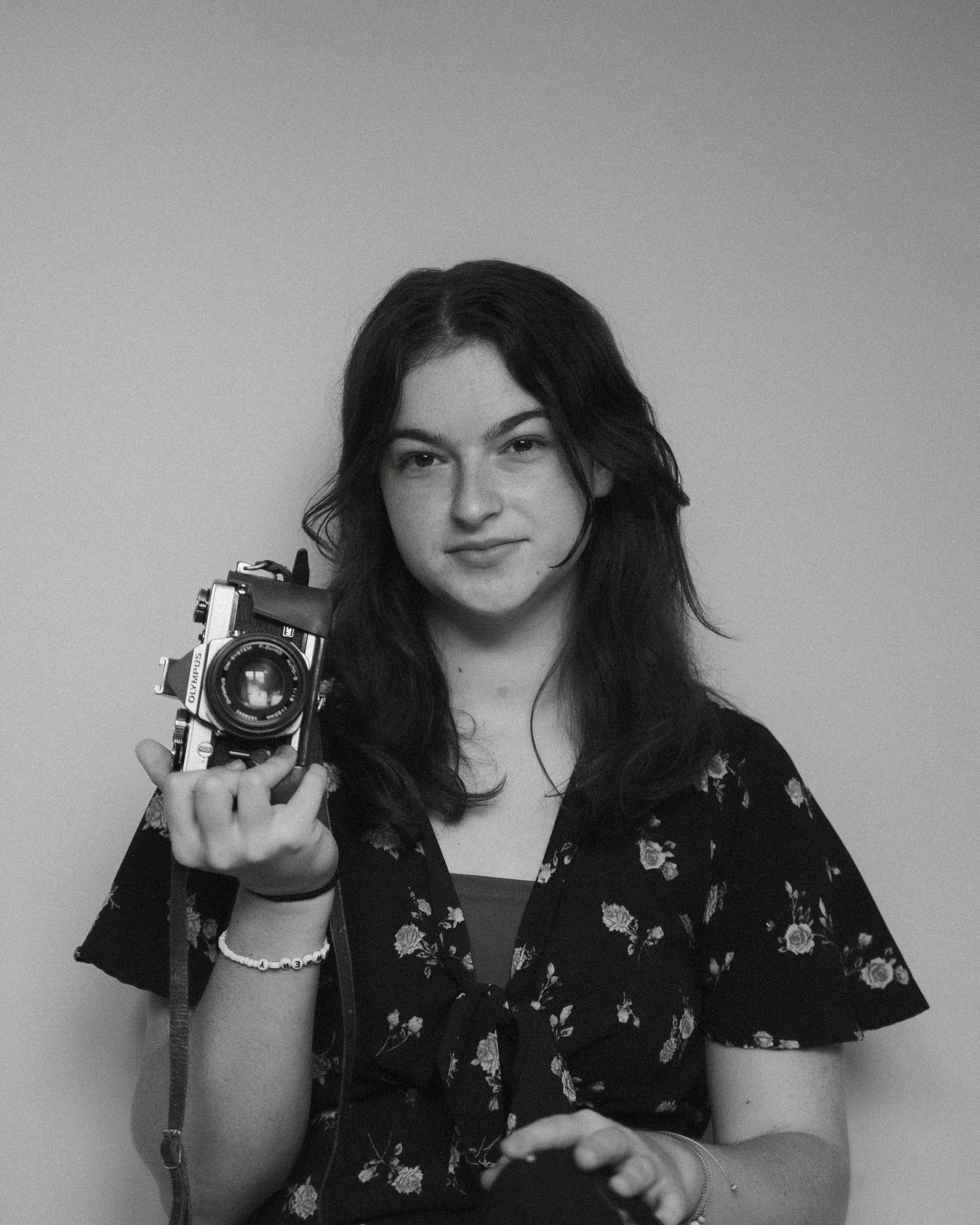 Black and white portrait of young woman holding a vintage film camera against a light gray background.