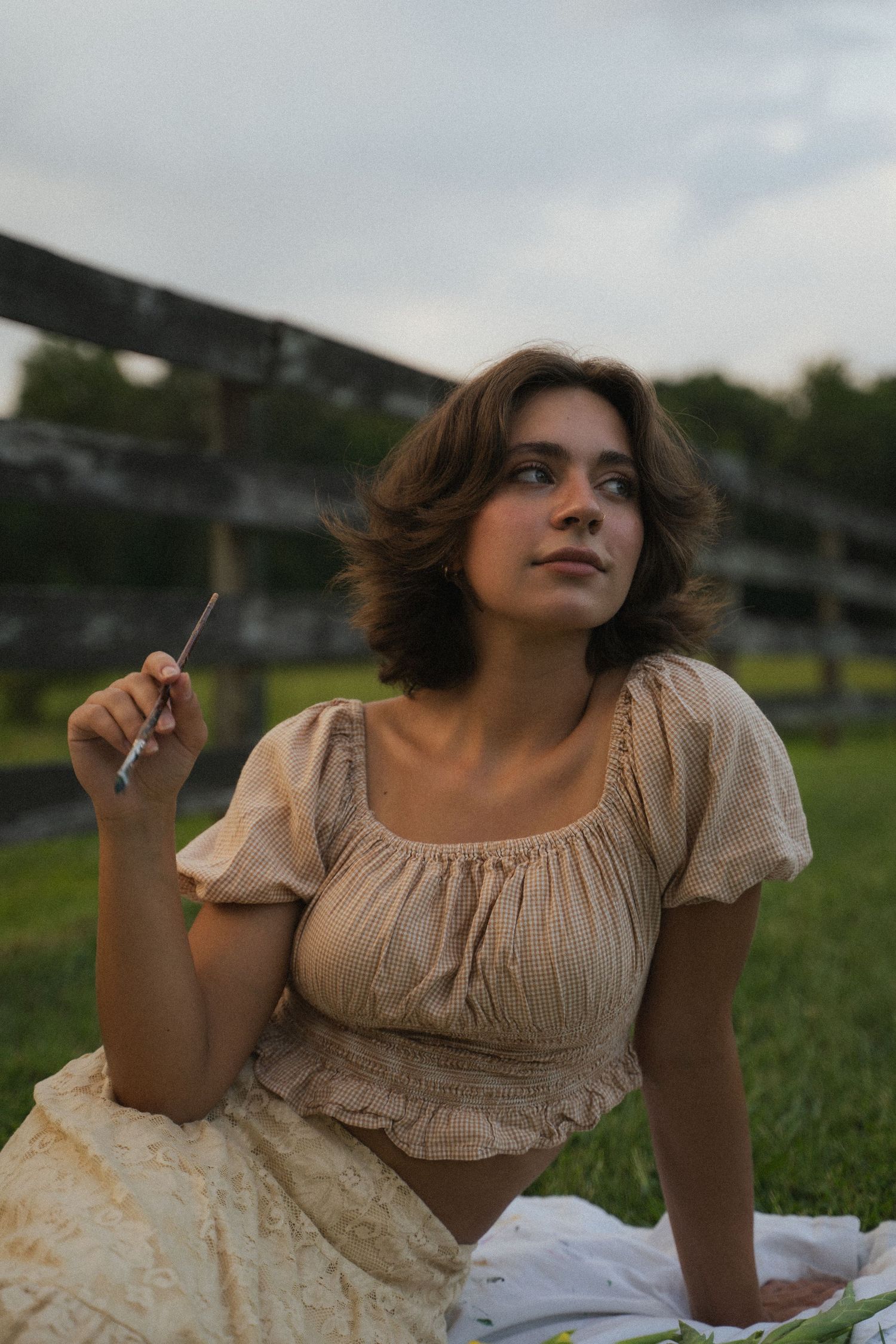 Young woman in vintage beige blouse sitting on grass holding paintbrush in open field.