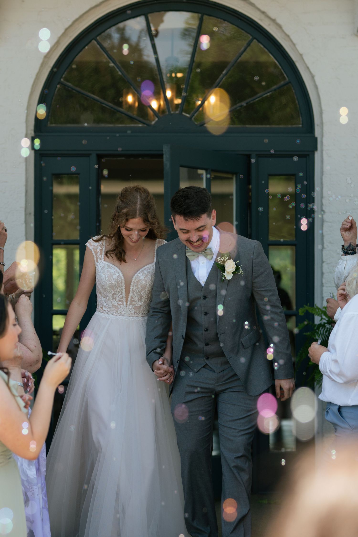 Bride and groom exit venue holding hands as colorful bubbles float around them in celebration.