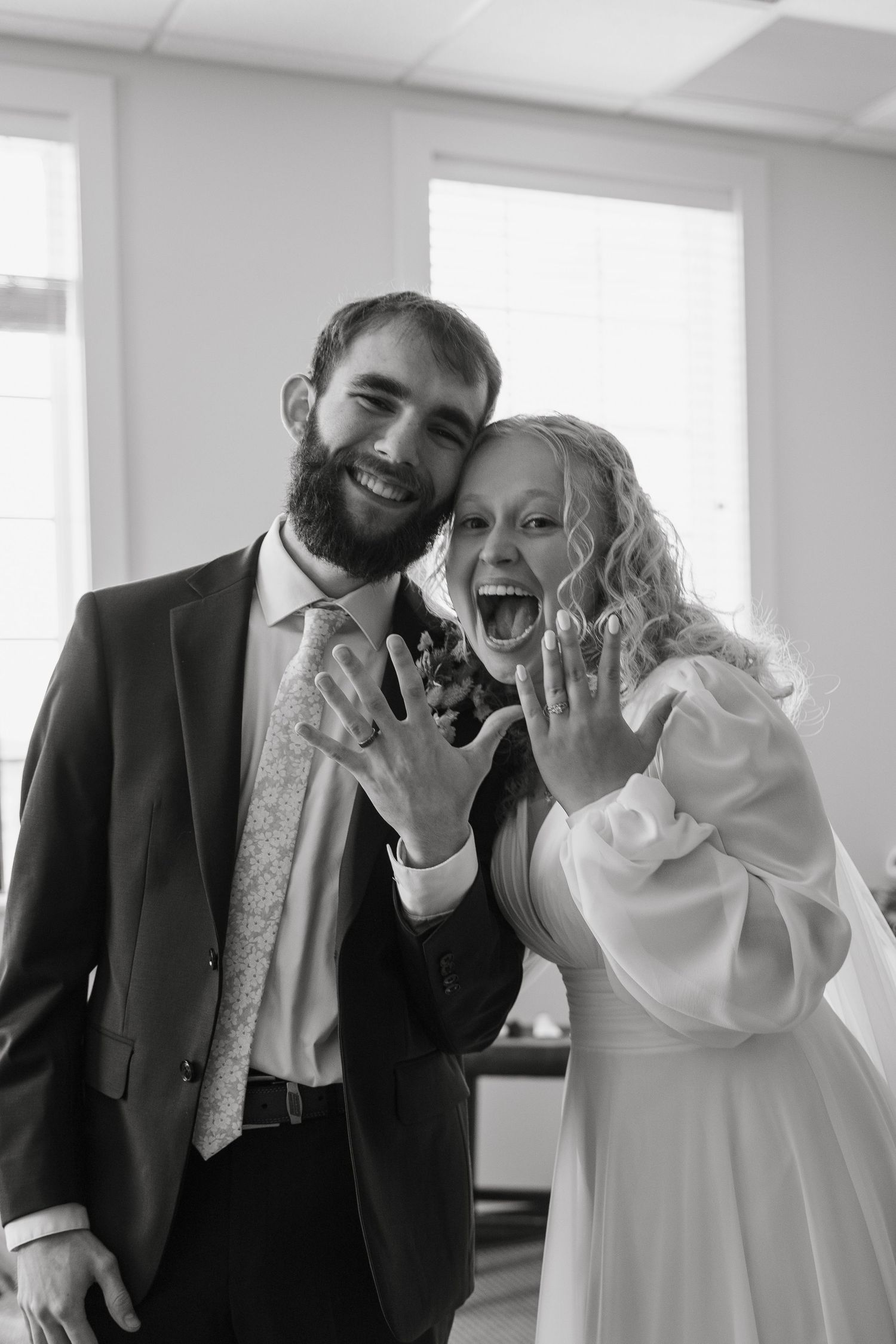 Black and white photo of newlyweds joyfully showing off wedding rings, bride in white dress, groom in suit.