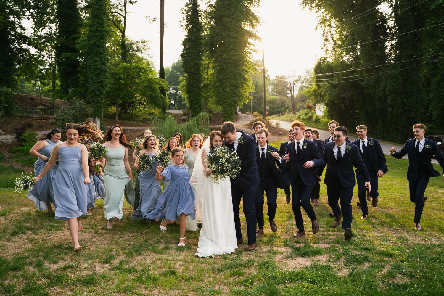 Wedding party walking joyfully outdoors in summer greenery, bridesmaids in blue dresses and groomsmen in suits.