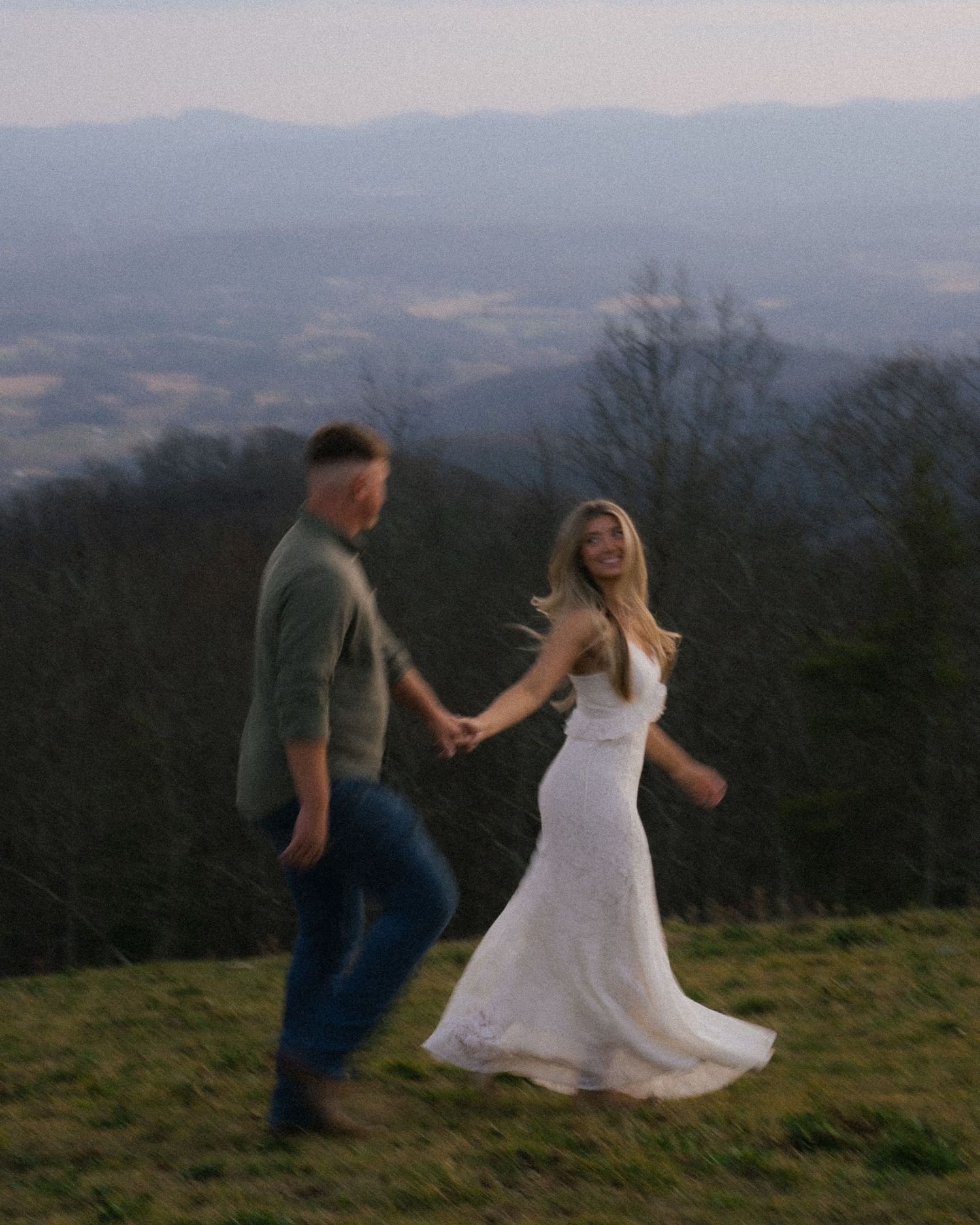 Couple holding hands walking on hilltop in white dress with scenic mountain valley view.