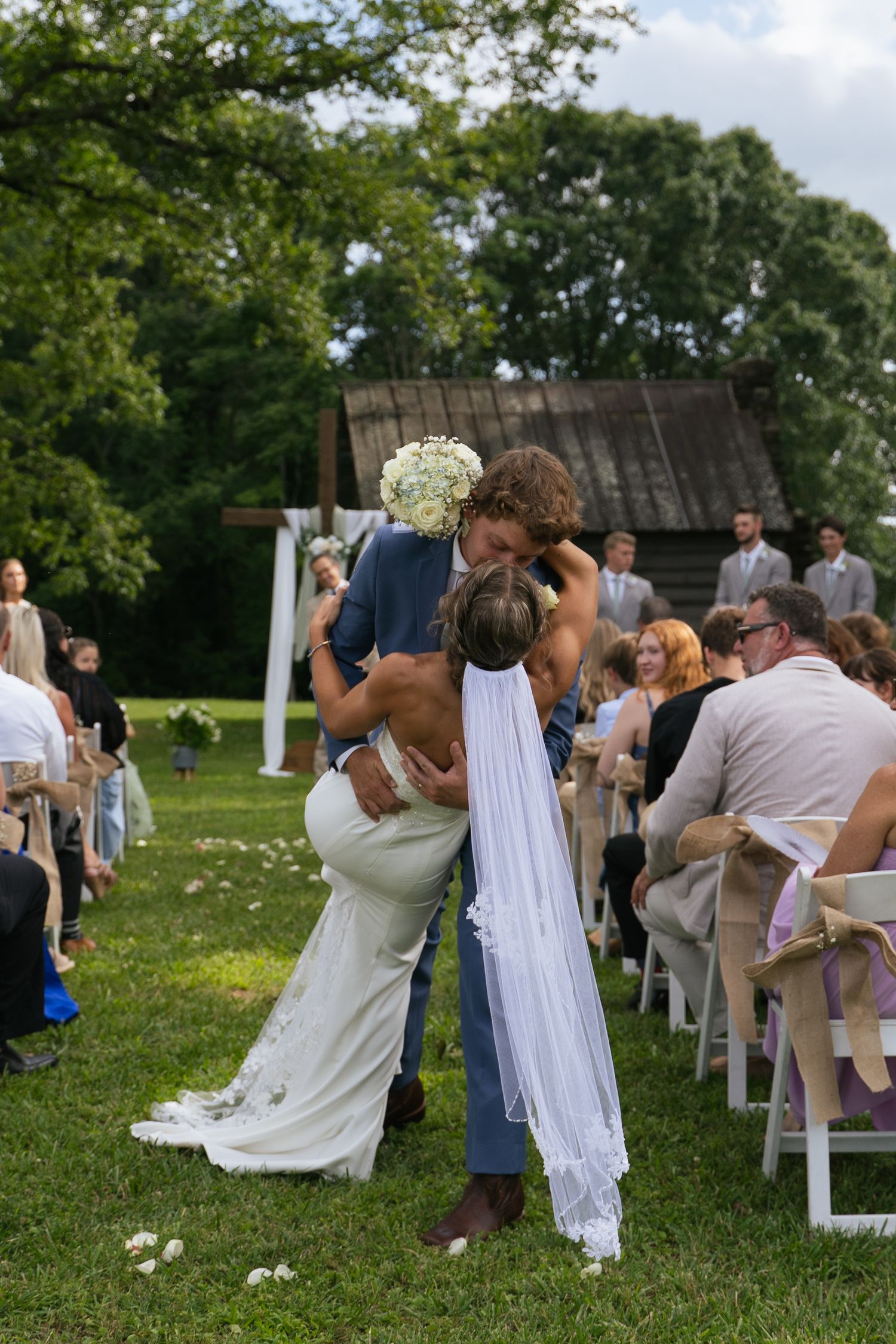 Groom dips bride for romantic kiss at outdoor wedding ceremony while guests watch from white chairs on green lawn.
