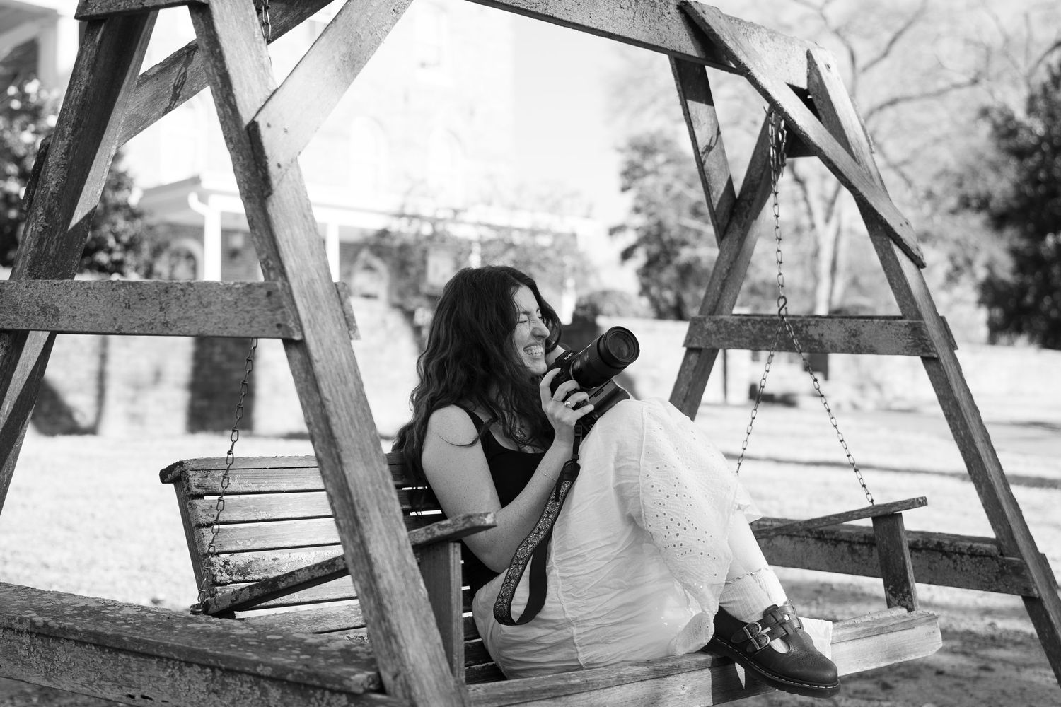 Black and white photo of woman with camera on wooden swing set, smiling.