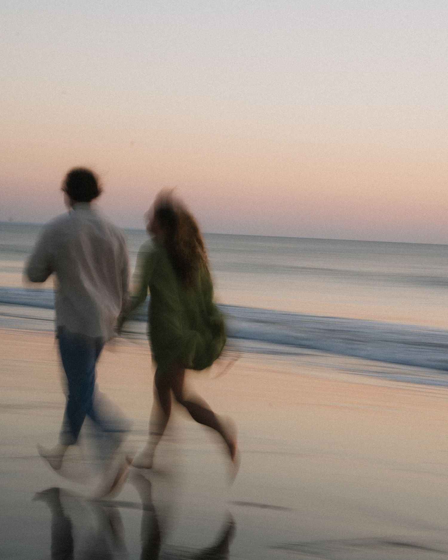 Blurred motion shot of couple holding hands running on beach at sunset with pink sky.