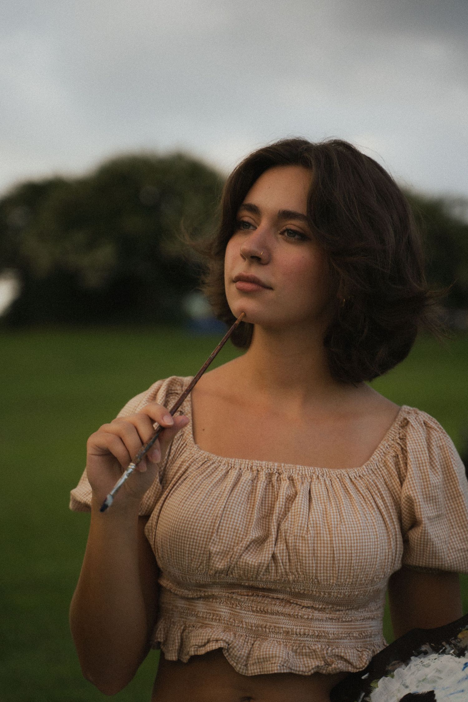 Young brunette woman in beige crop top holding paintbrush outdoors on cloudy day.