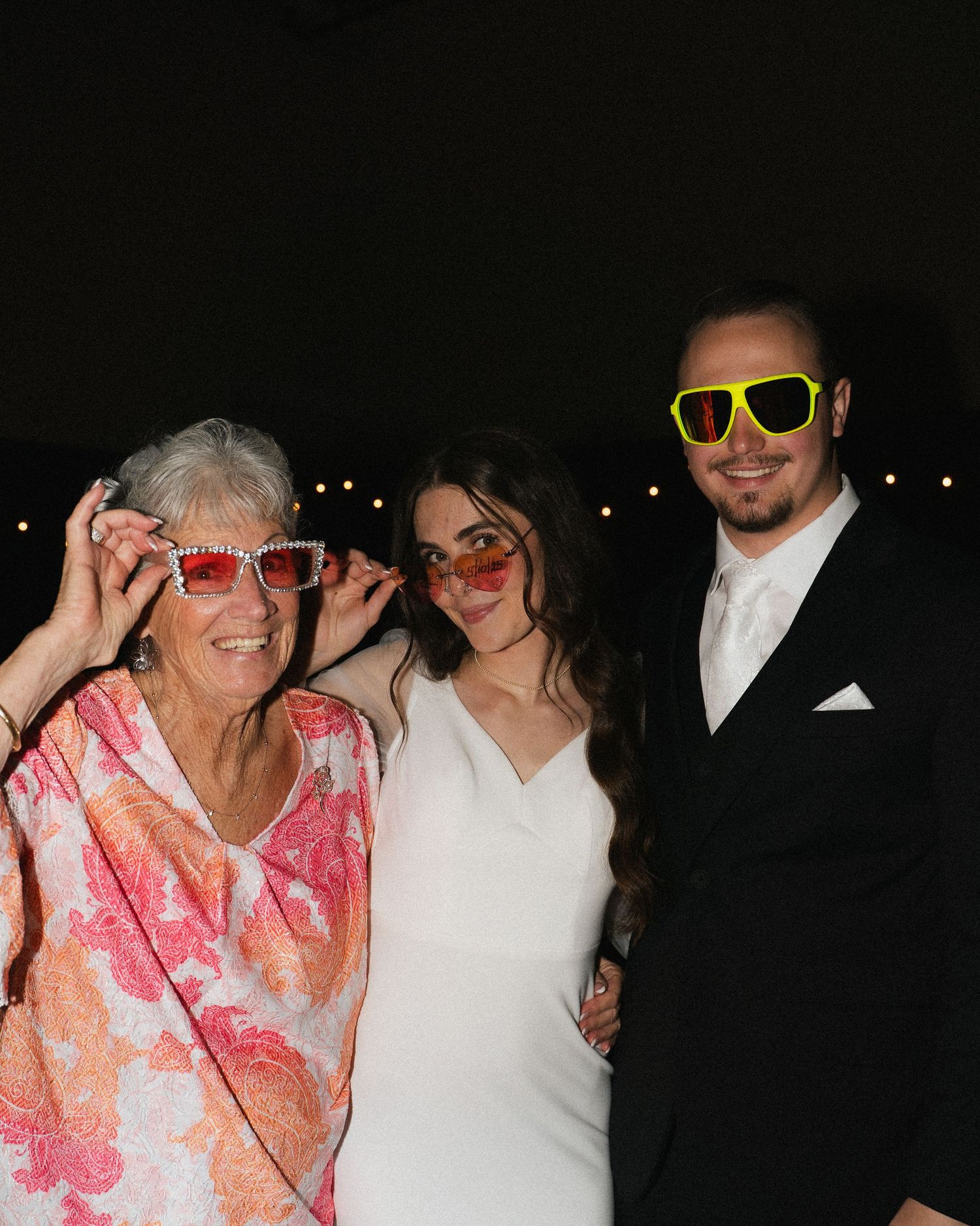 Bride, groom, and elderly woman posing playfully with oversized colorful sunglasses at a wedding reception.