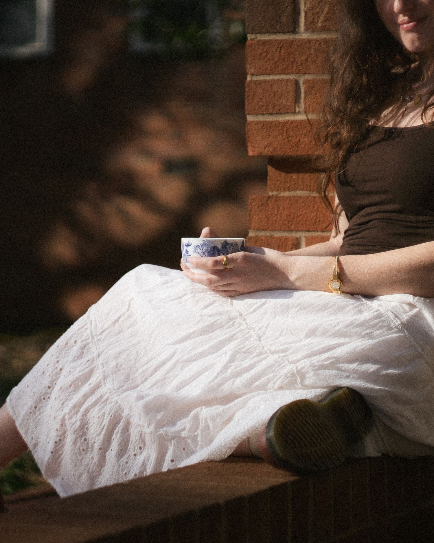 Woman in white eyelet skirt holding blue and white ceramic cup while sitting on brick steps.