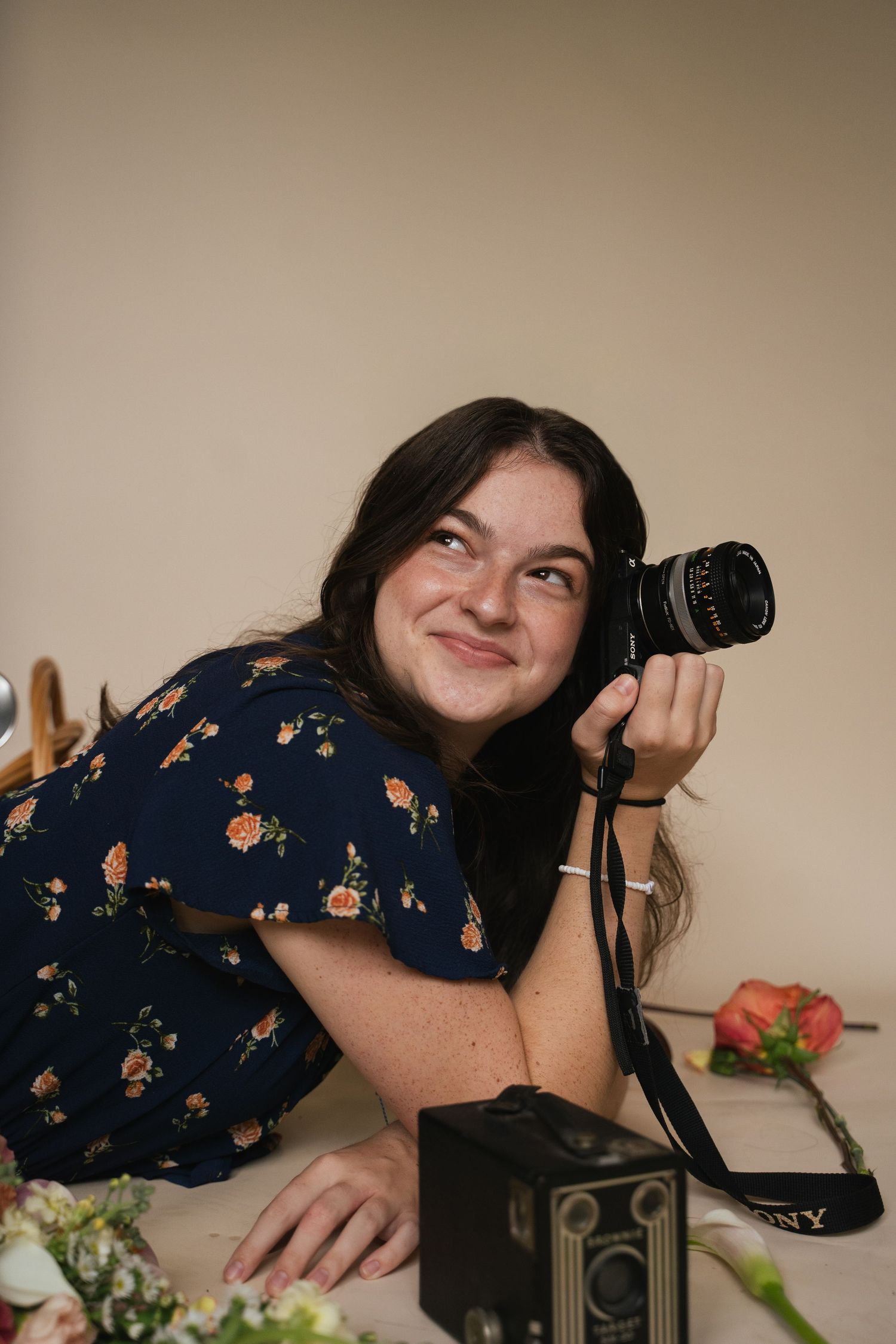 Smiling young woman in floral dress holding Sony camera while lying on surface with flowers.
