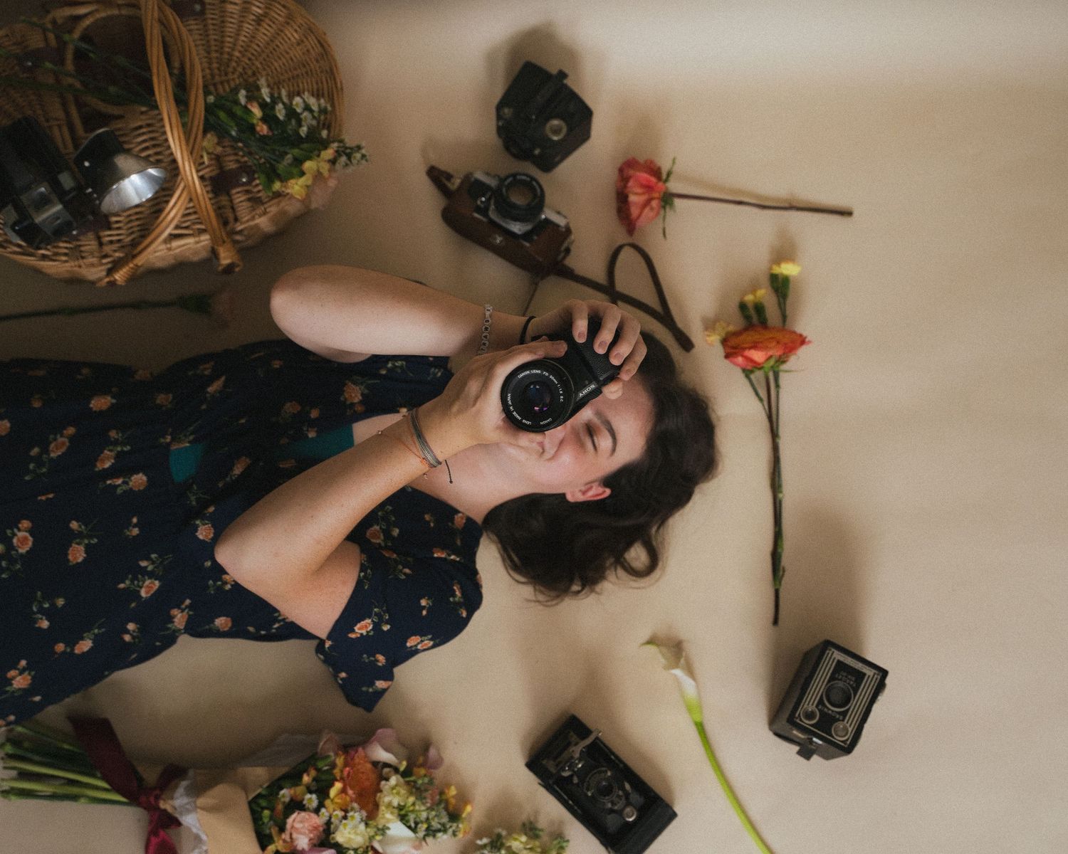 Woman lying on floor surrounded by vintage cameras and flowers, holding camera to her face in artistic flat lay photo.