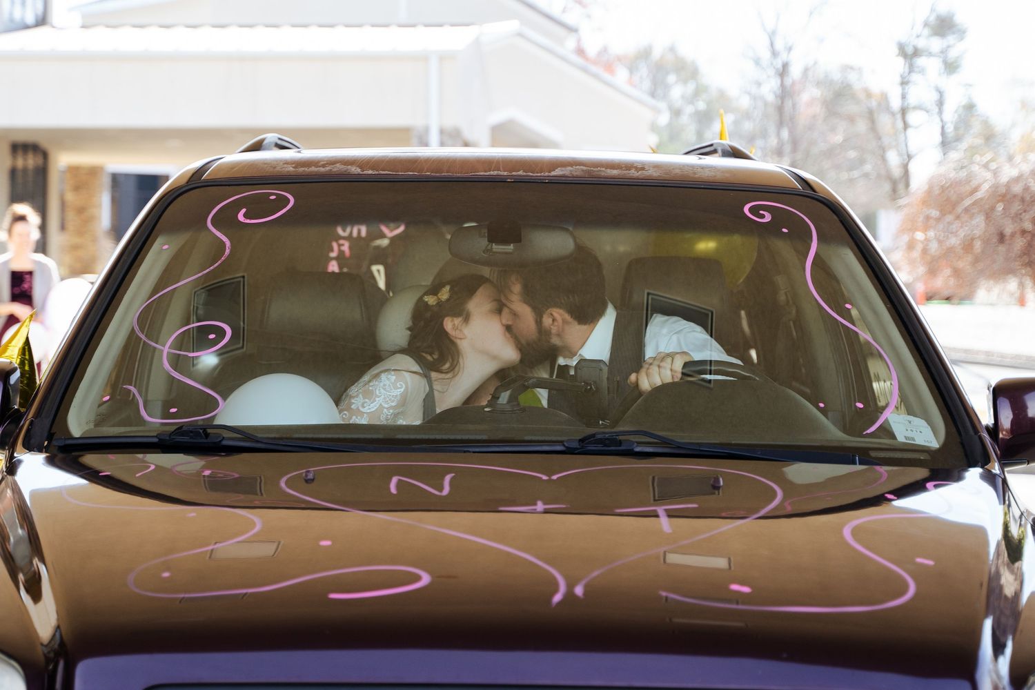Newlyweds sharing a kiss inside a decorated getaway car with wedding messages written on the windshield.