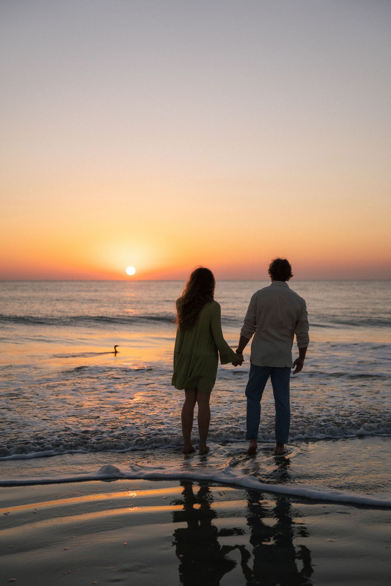 Couple holding hands watching vibrant ocean sunset with silhouetted bird in background.