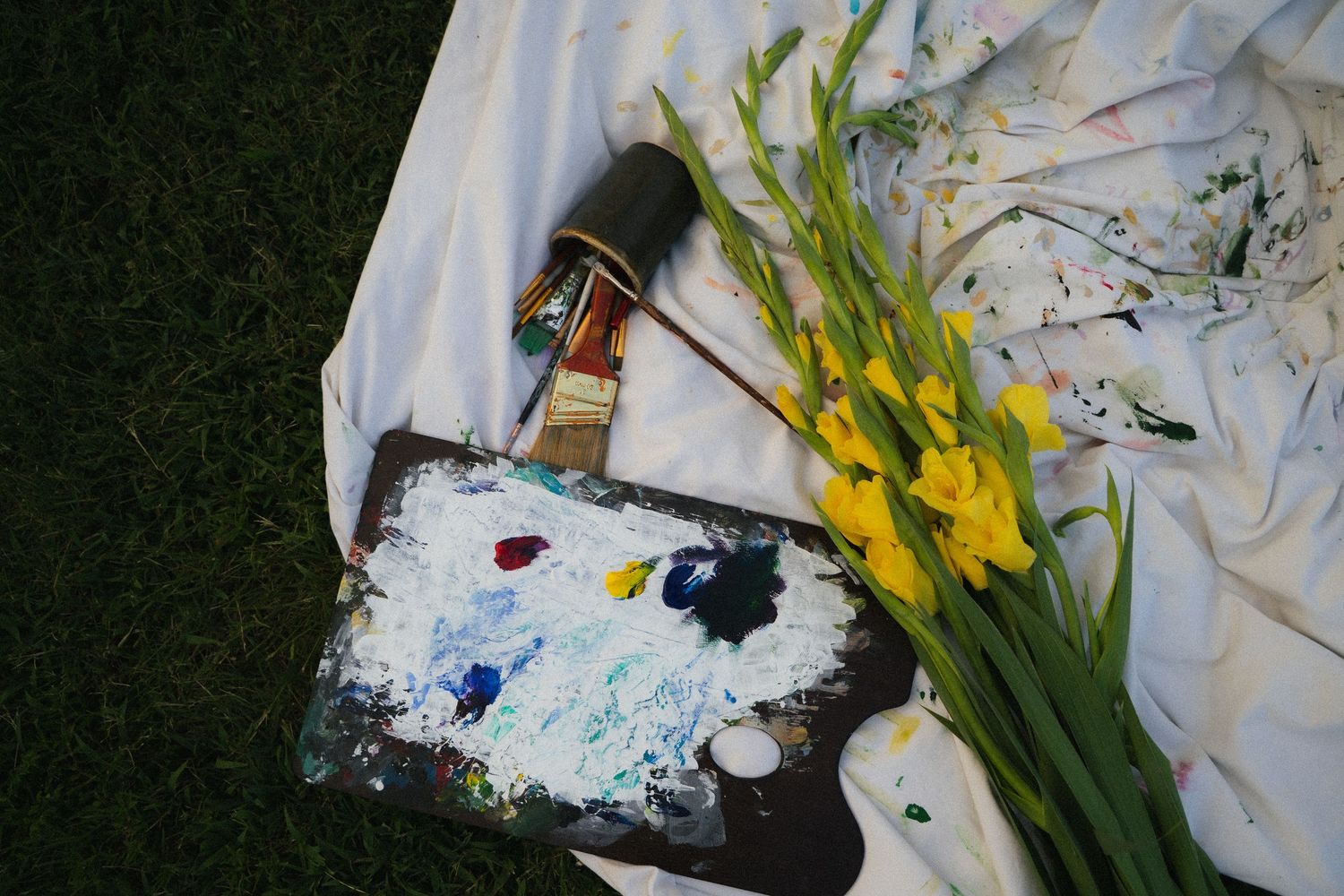Artist palette with paint, paintbrush, and yellow gladiolus flowers on a white cloth outdoors.