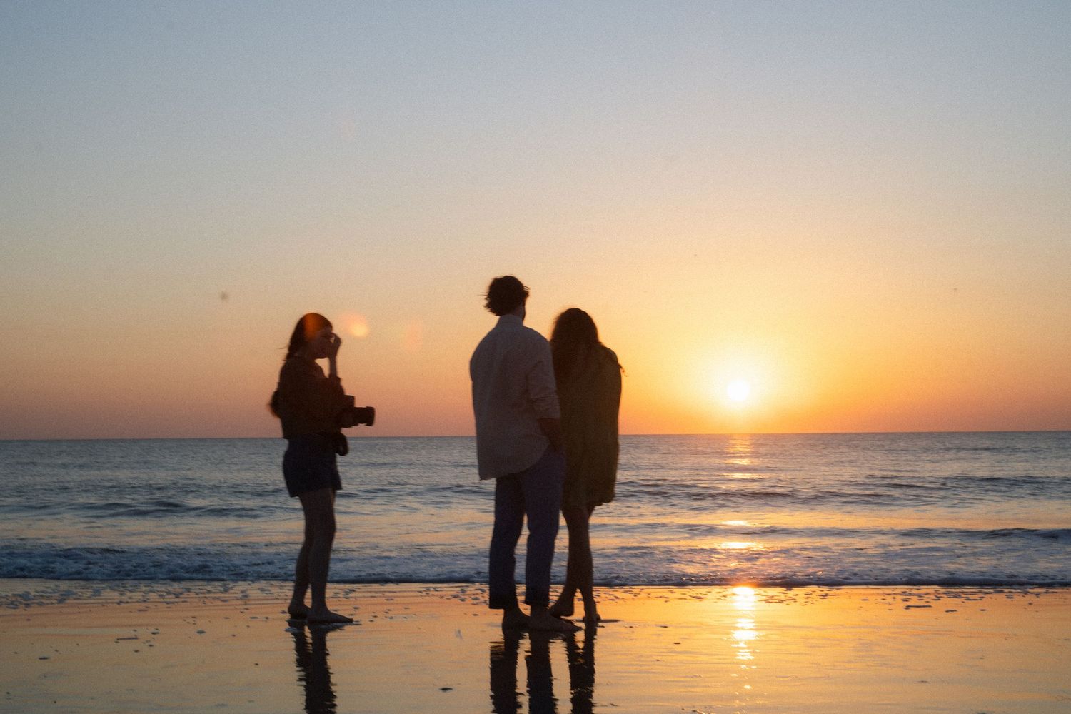 Three people silhouetted on a beach at sunset, one holding a camera, capturing a romantic golden hour moment.