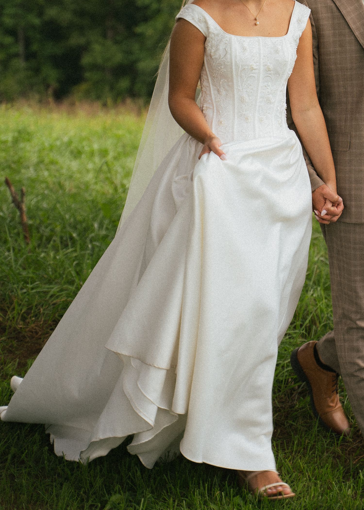 Close-up of bride in elegant white corset bodice wedding gown with layered skirt holding grooms hand outdoors.