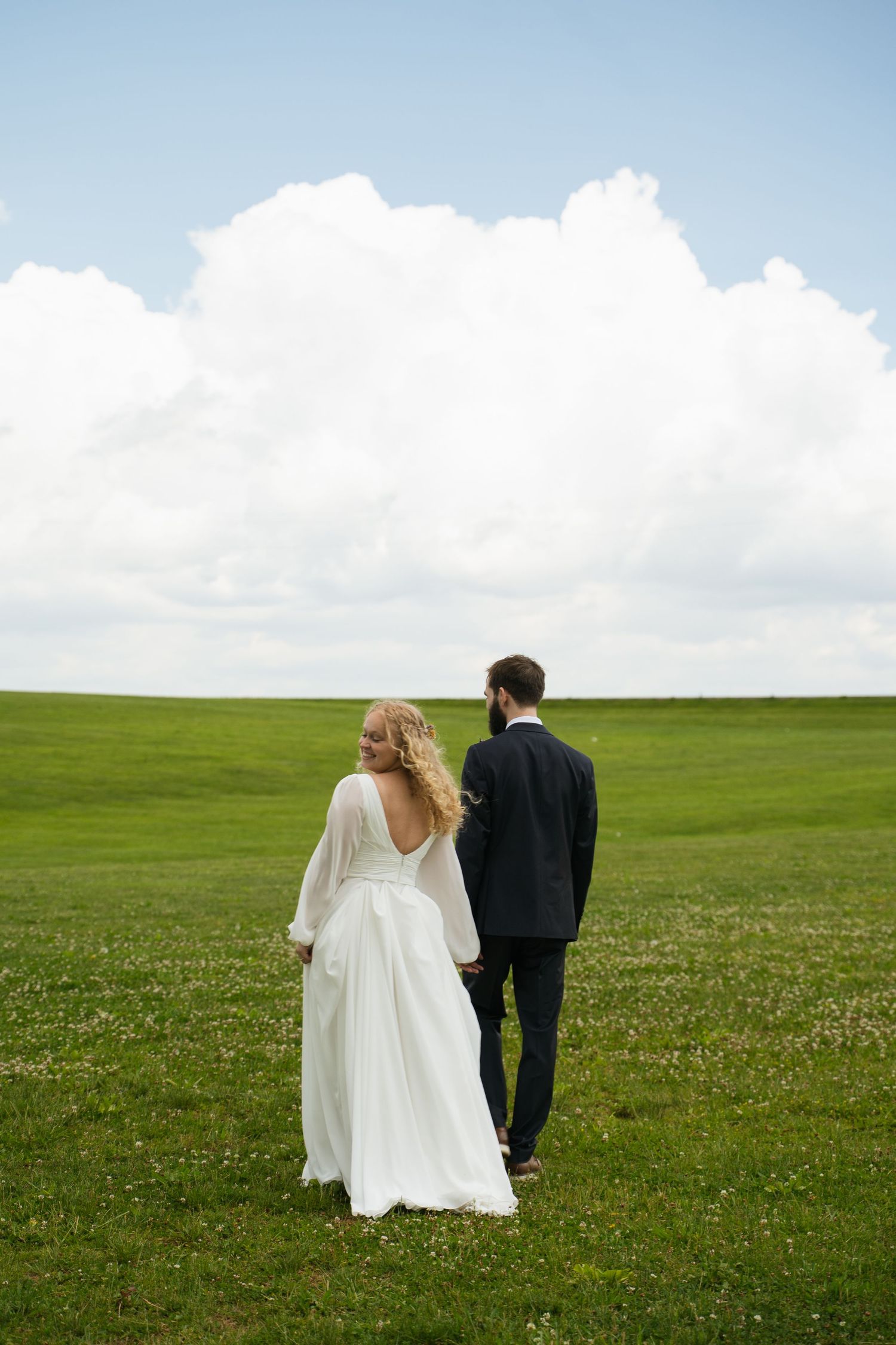 Bride and groom walk hand-in-hand through vast green meadow under dramatic white clouds in open sky.