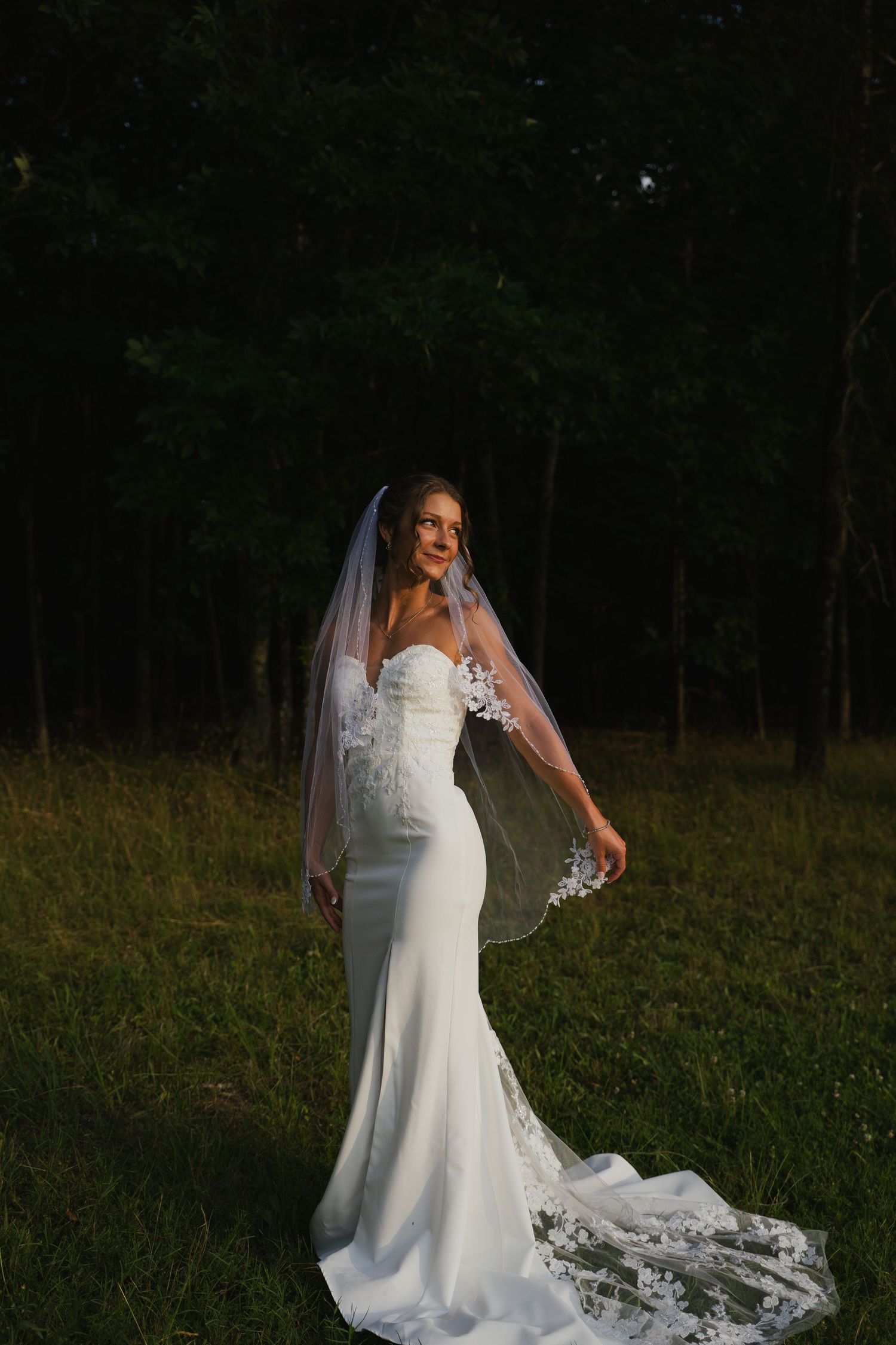Elegant bride in fitted lace wedding gown and veil posing in golden hour light against dark forest.
