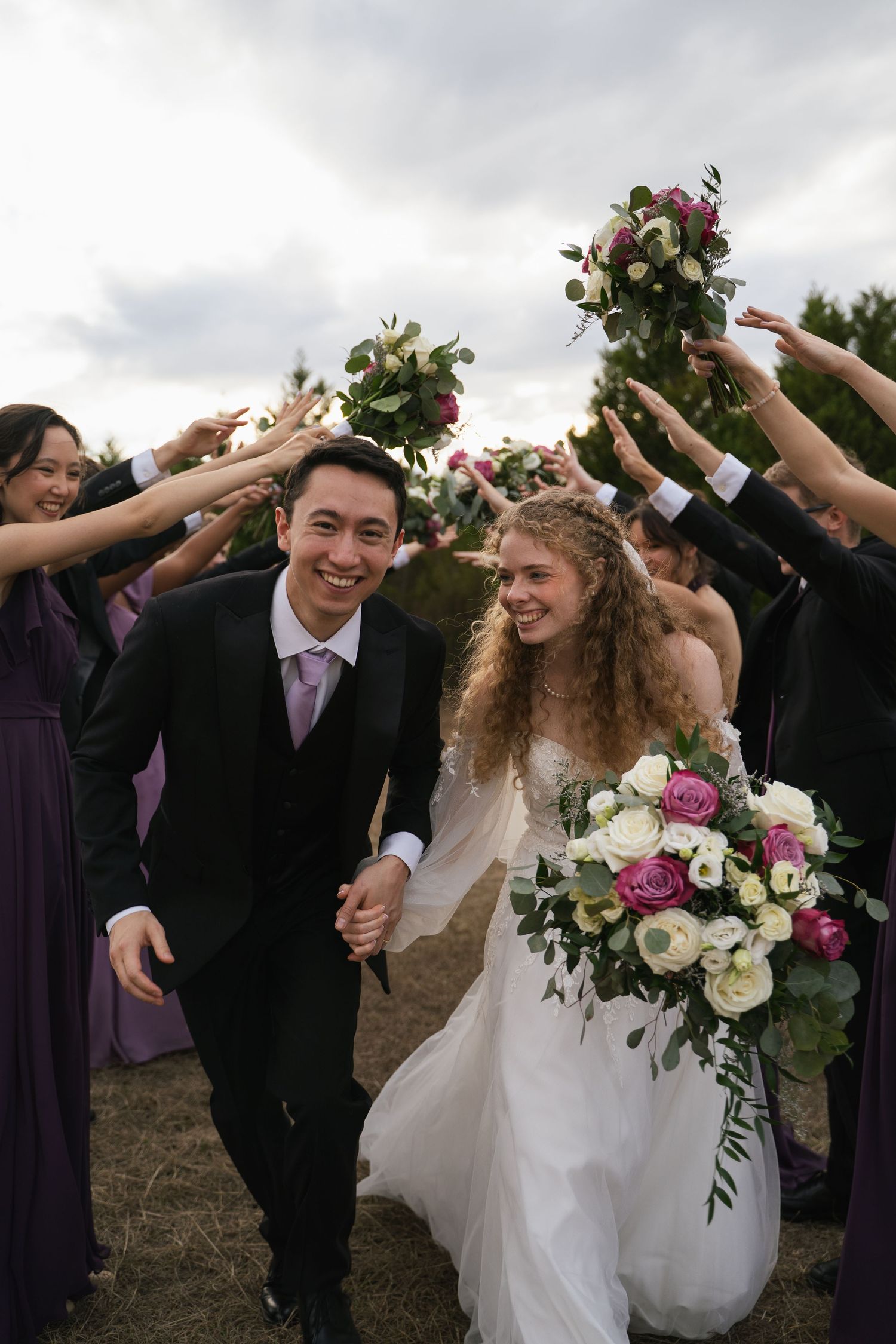 Joyful newlyweds walk hand-in-hand under an arch of raised bouquets held by bridesmaids and groomsmen outdoors.